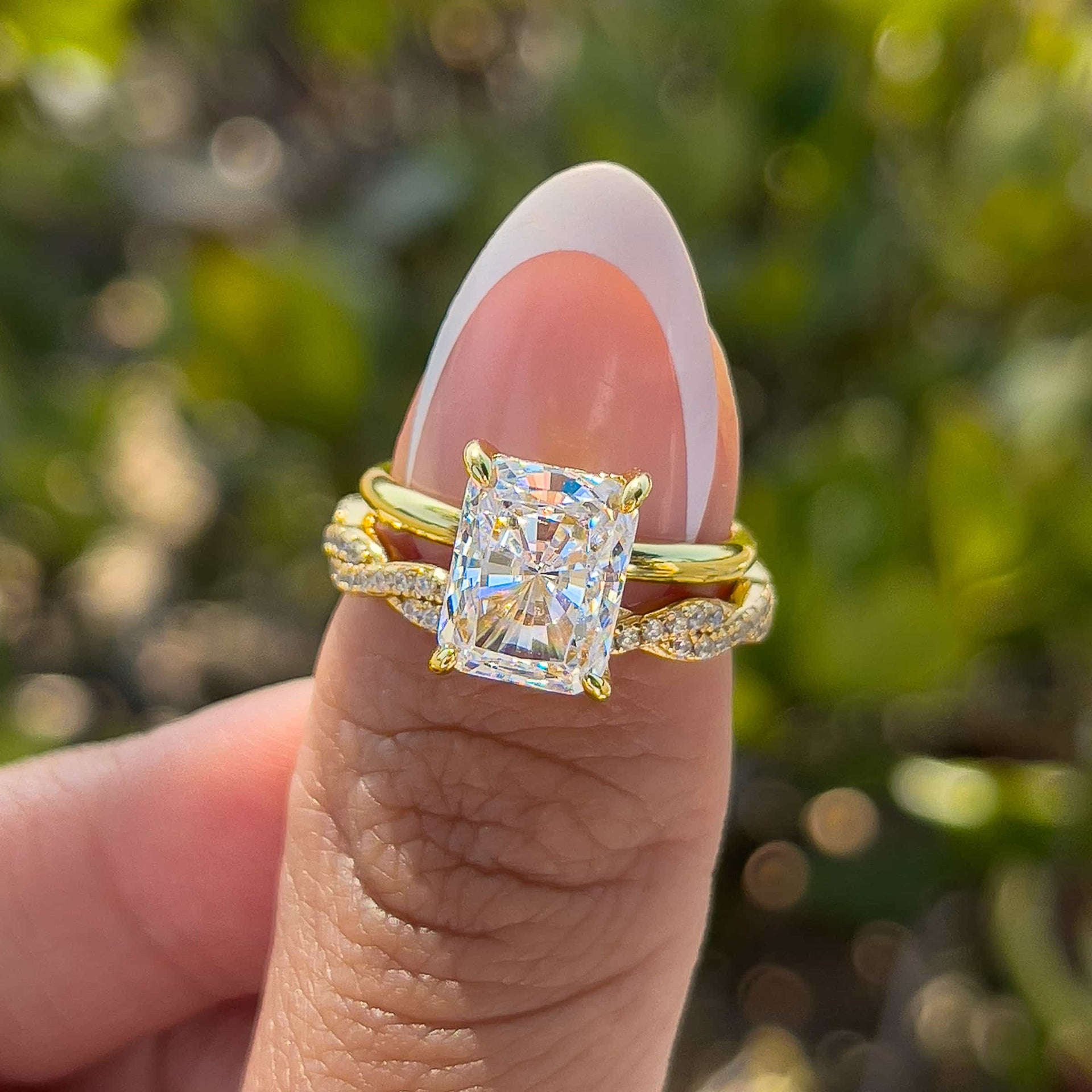 gold 3 carat radiant cut solitaire and twisting eternity wedding band paired together to create a timeless, yet unique wedding set on model with french tip nails in front of greenery