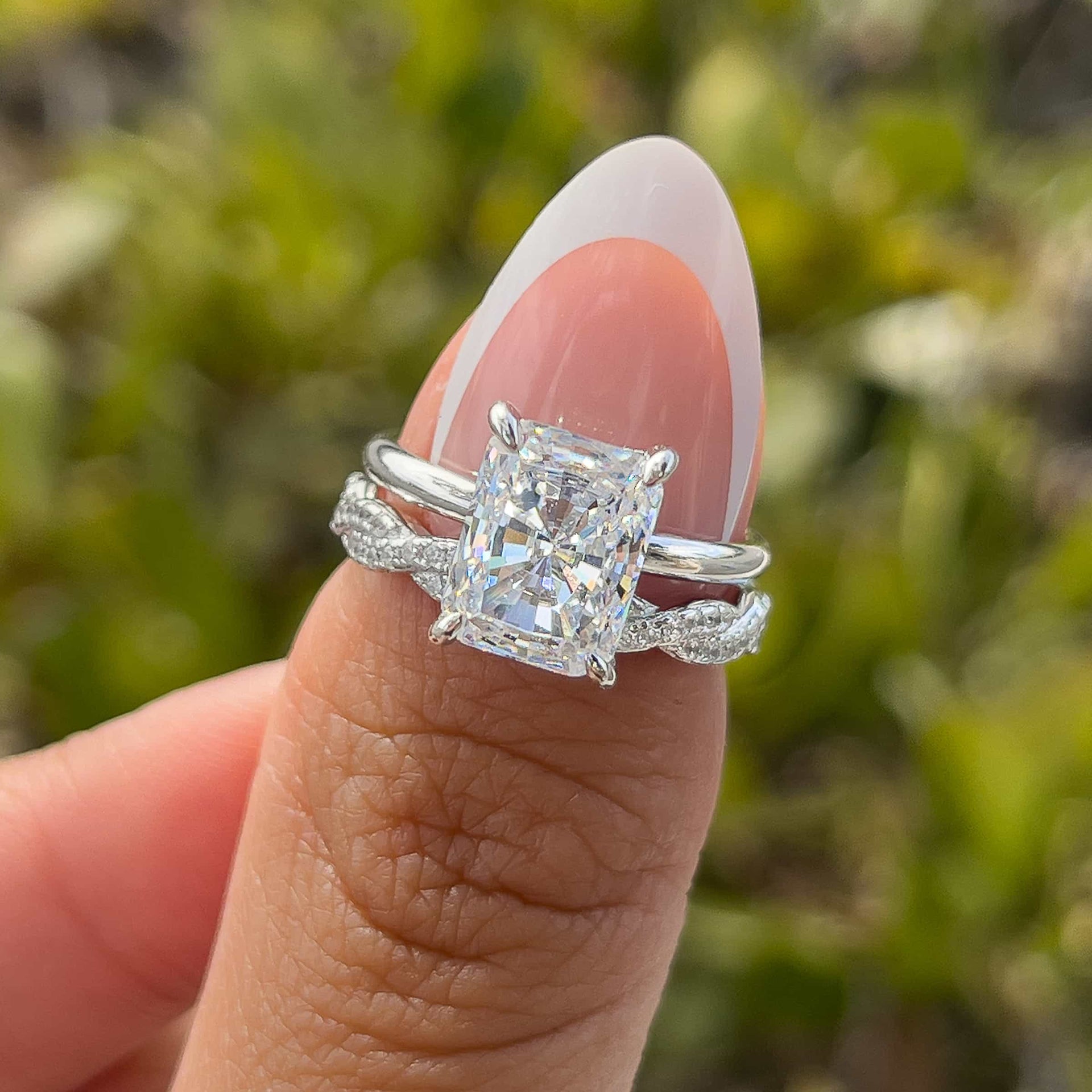 silver 3 carat radiant cut solitaire and twisting eternity wedding band paired together to create a timeless, yet unique wedding set on model with french tip nails in front of greenery