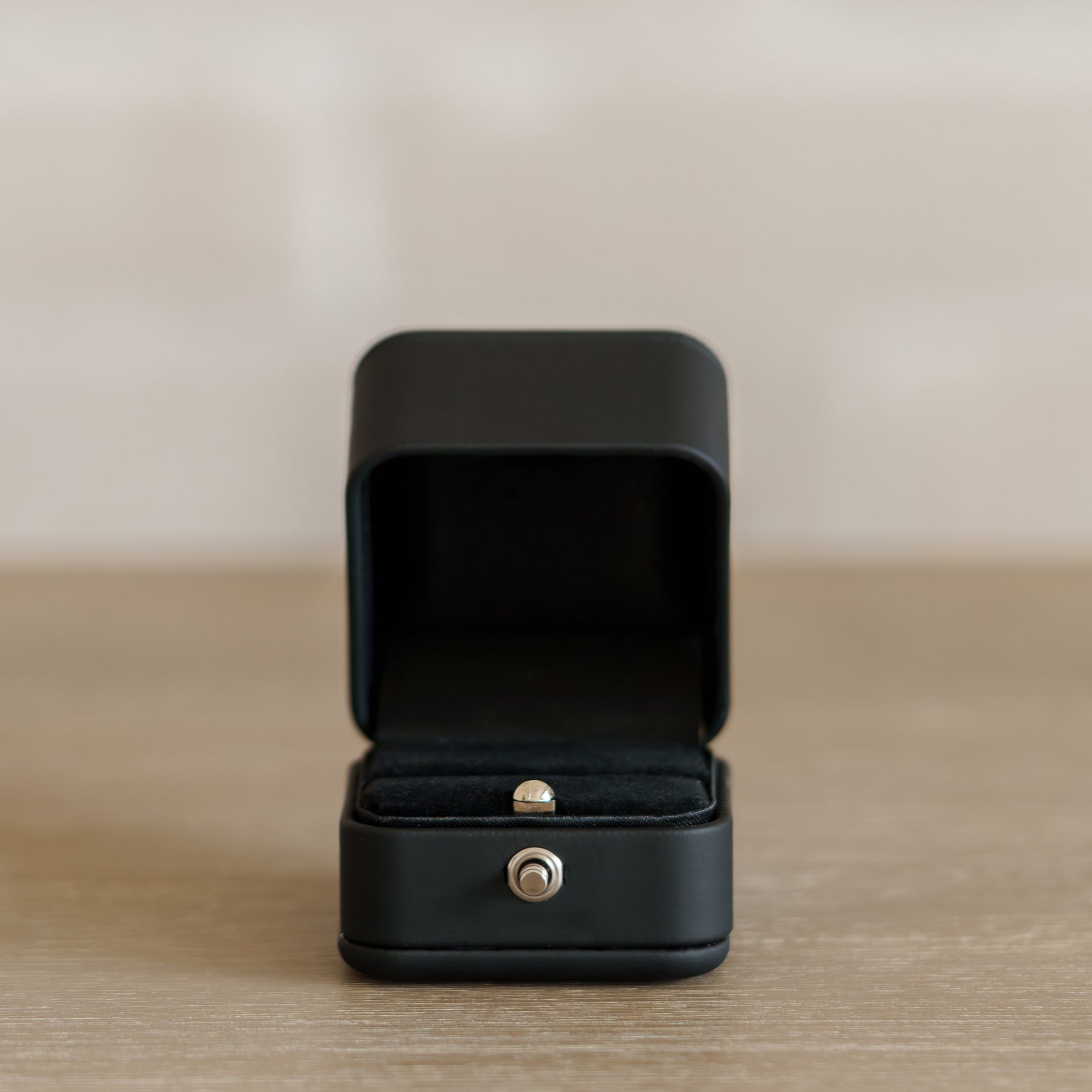 simple, matte black ring box with silver push button in an open position on a wooden surface