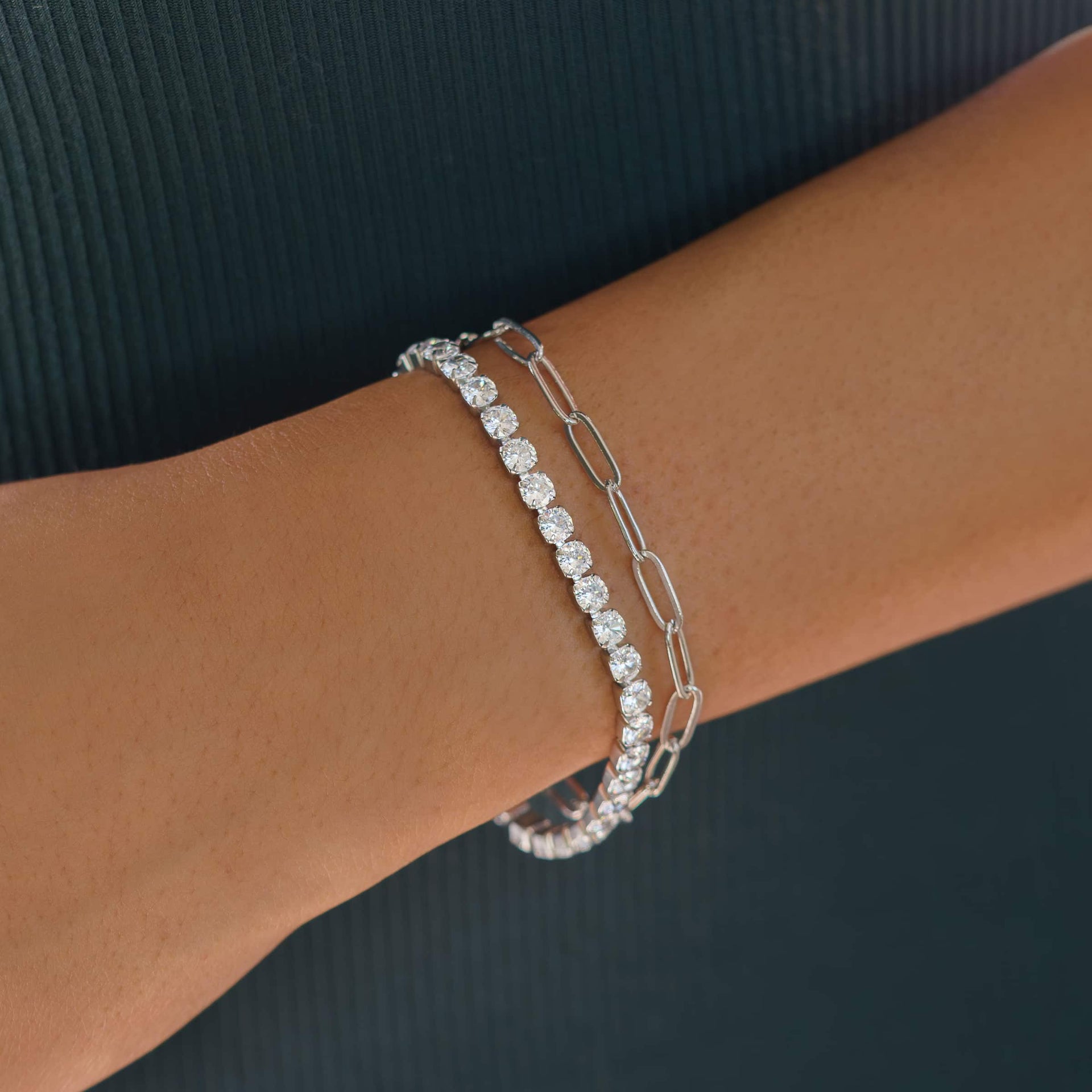 everyday silver tennis bracelet worn as a part of a bracelet stack with a silver paperclip necklace by model wearing dark colors
