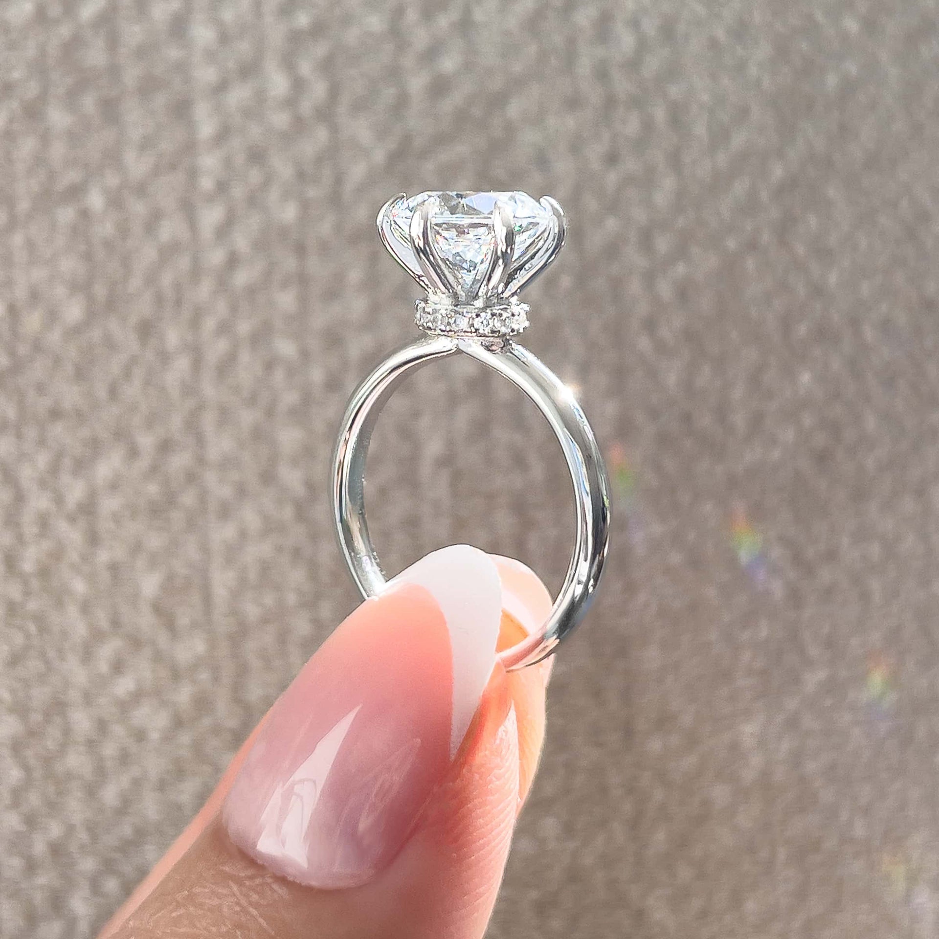 Side view of silver 6 prong 2 carat round cut solitaire engagement ring with secret halo detail held by model with french tip nails