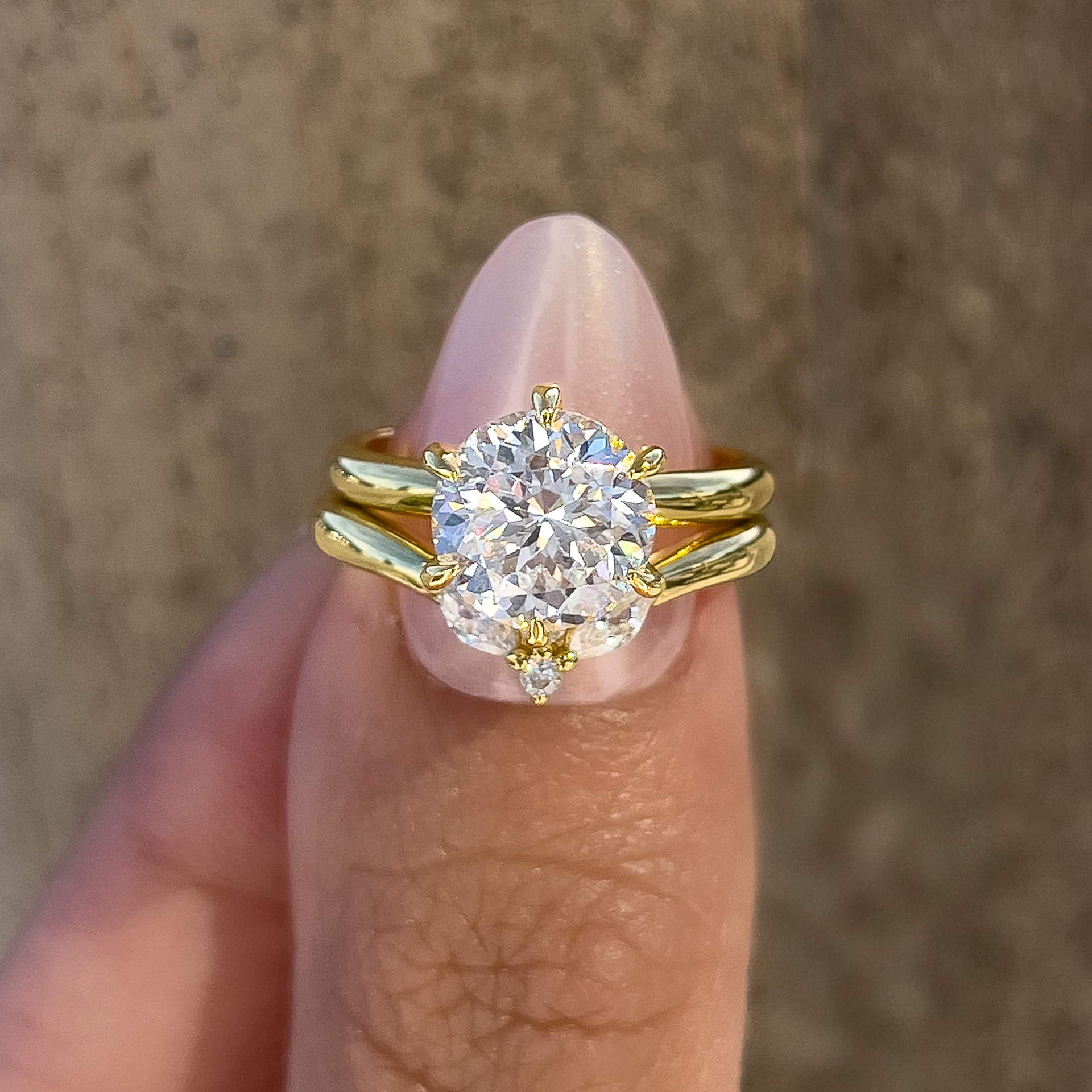 Gold wedding ring set made up of a 2 carat round cut solitaire engagement ring and dainty chevron wedding band, held together by a model with light pink nails