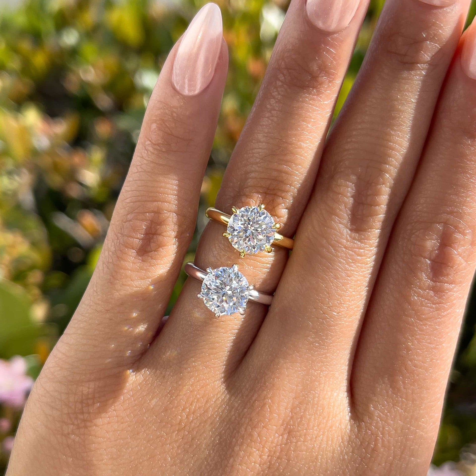 A 2 carat round cut, 6 prong solitaire engagement ring shown in gold and silver on model with chrome light pink nails
