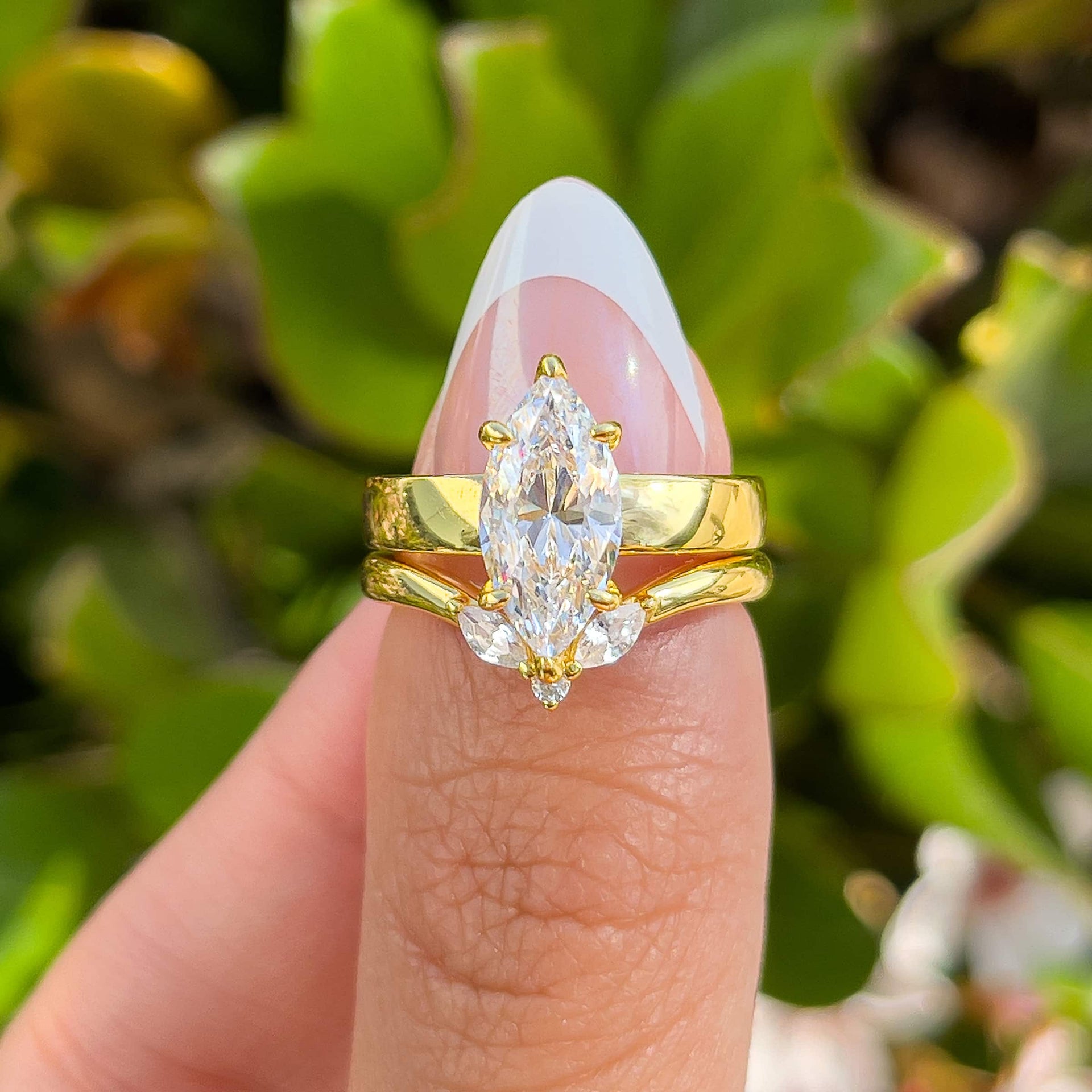 unique gold wedding ring set with a 2 carat marquise cut 3mm solitaire and a dainty chevron style band held by a model with french tip nails in front of greenery
