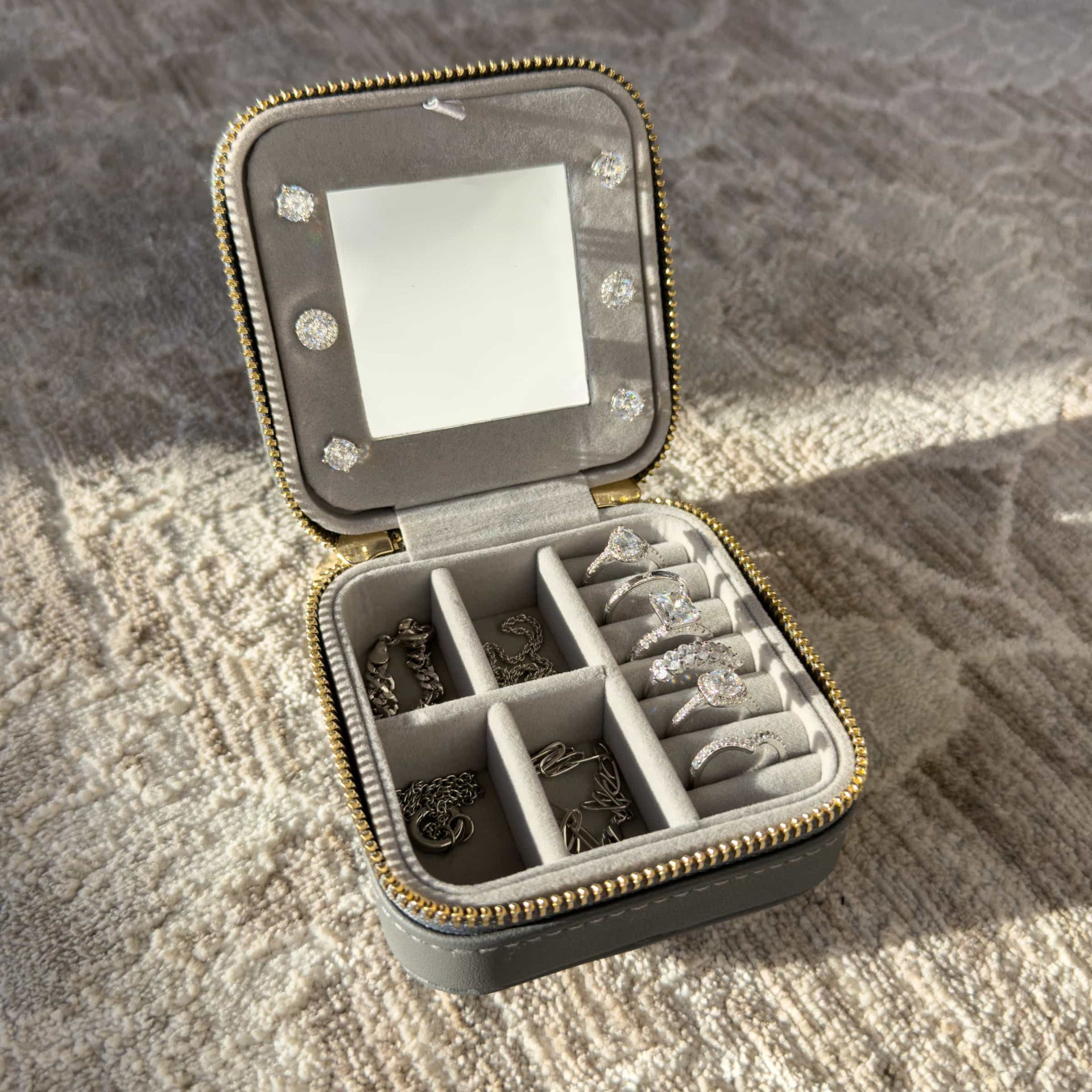 A charcoal color square shaped travel box with gold hardware opened to show the jewelry collection inside sitting on a neutral toned carpet