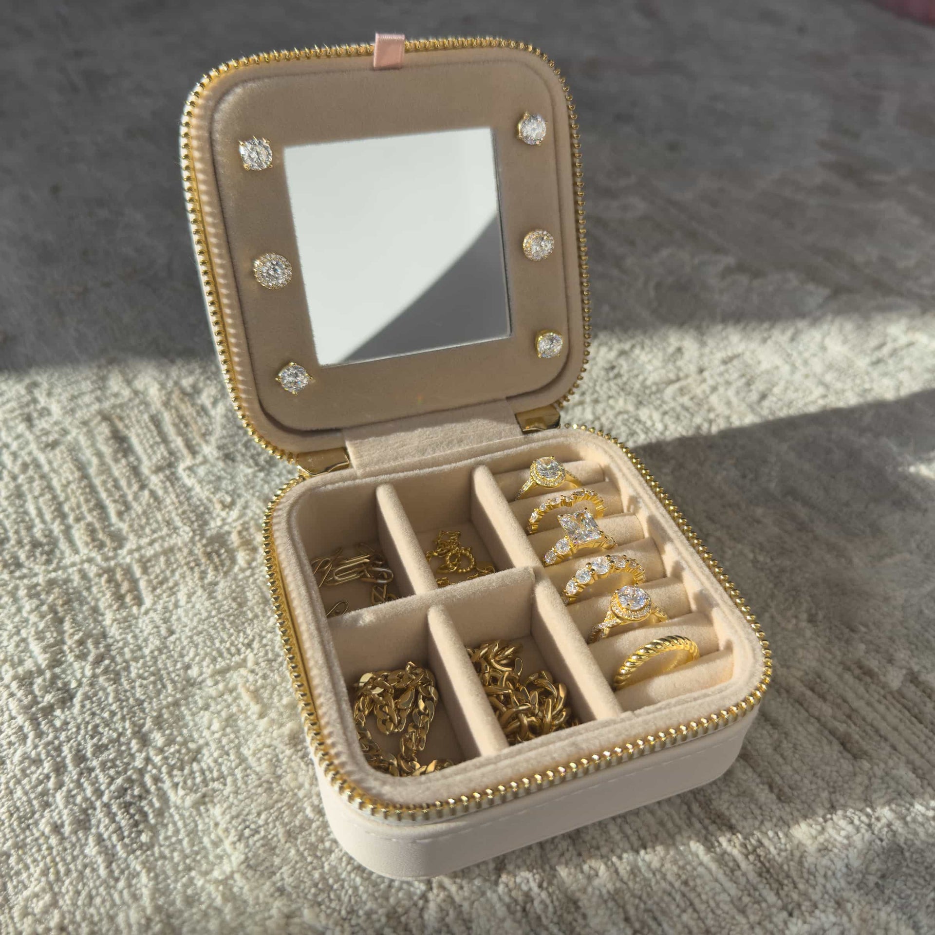 A cream color square shaped travel box with gold hardware opened to show the jewelry collection inside sitting on a neutral toned carpet