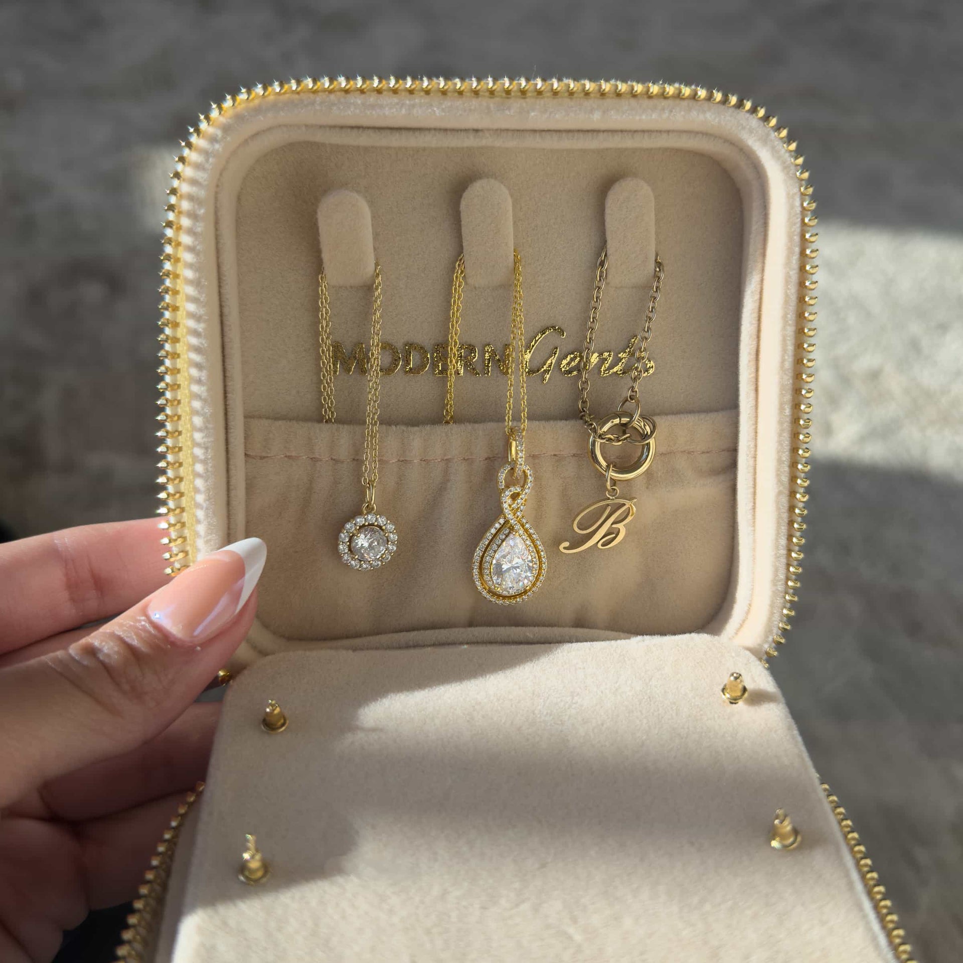 A cream color square shaped travel box with gold hardware opened from the top portion to showcase the hidden compartment for additional jewelry storage and the Modern Gents Logo. Held by a model with french tip nails