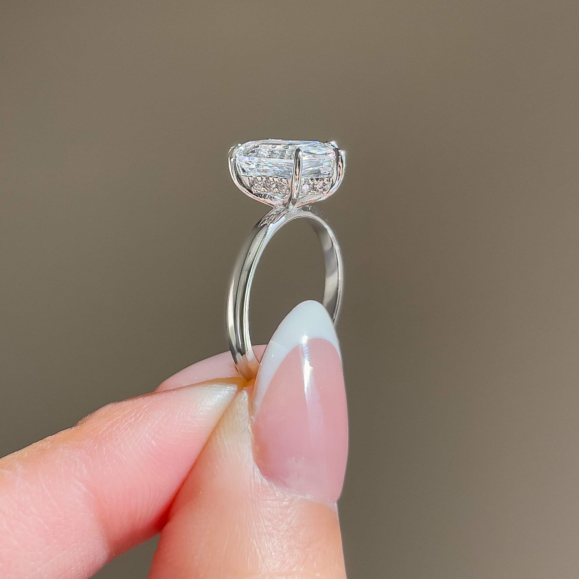 side view of a silver 2.5 carat hexagon cut solitaire hidden halo engagement ring held by a model with french tip nails