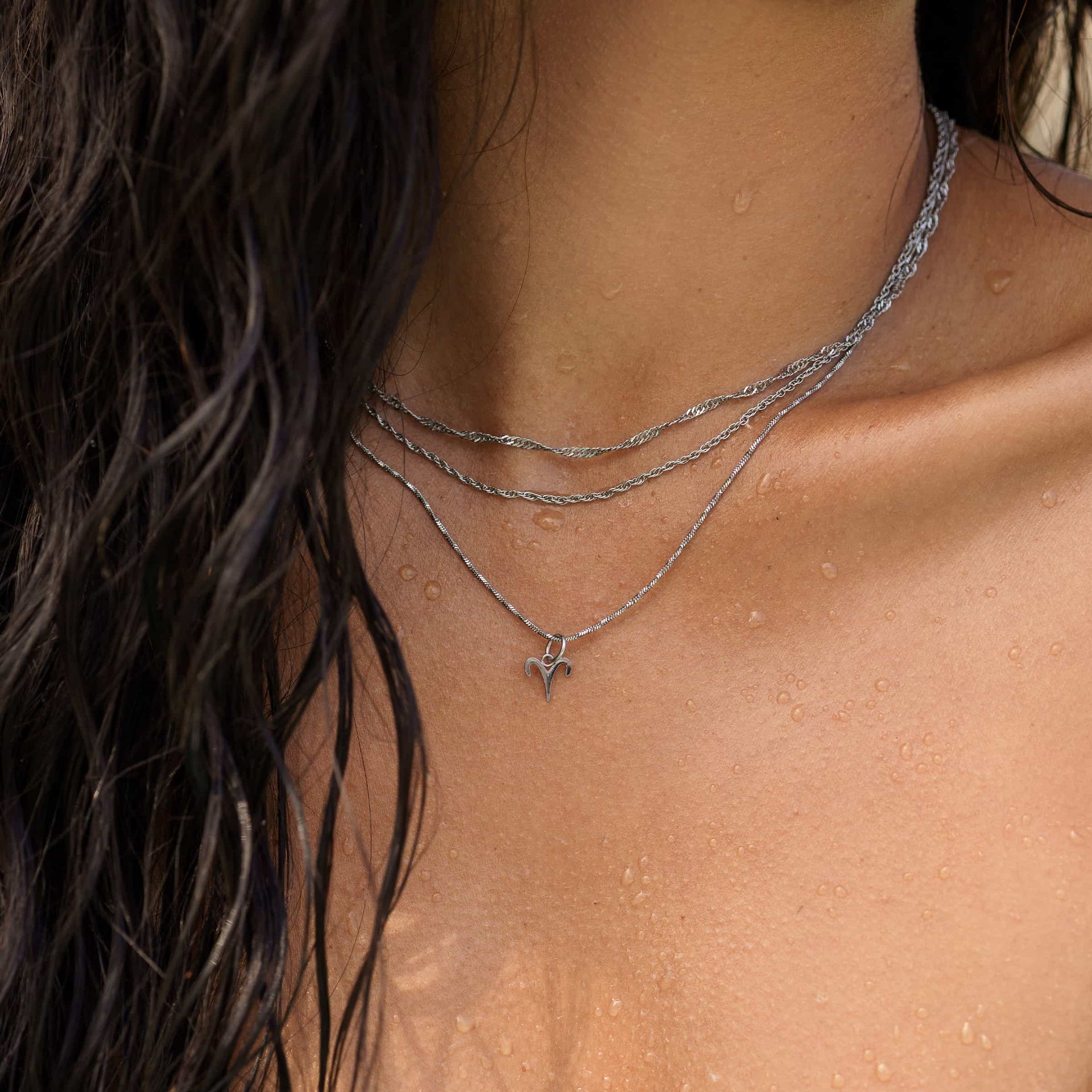 a close up of a model at the beach wearing a stainless steel twisted detail cable chain necklace layered with other dainty necklaces & a zodiac sign detail 