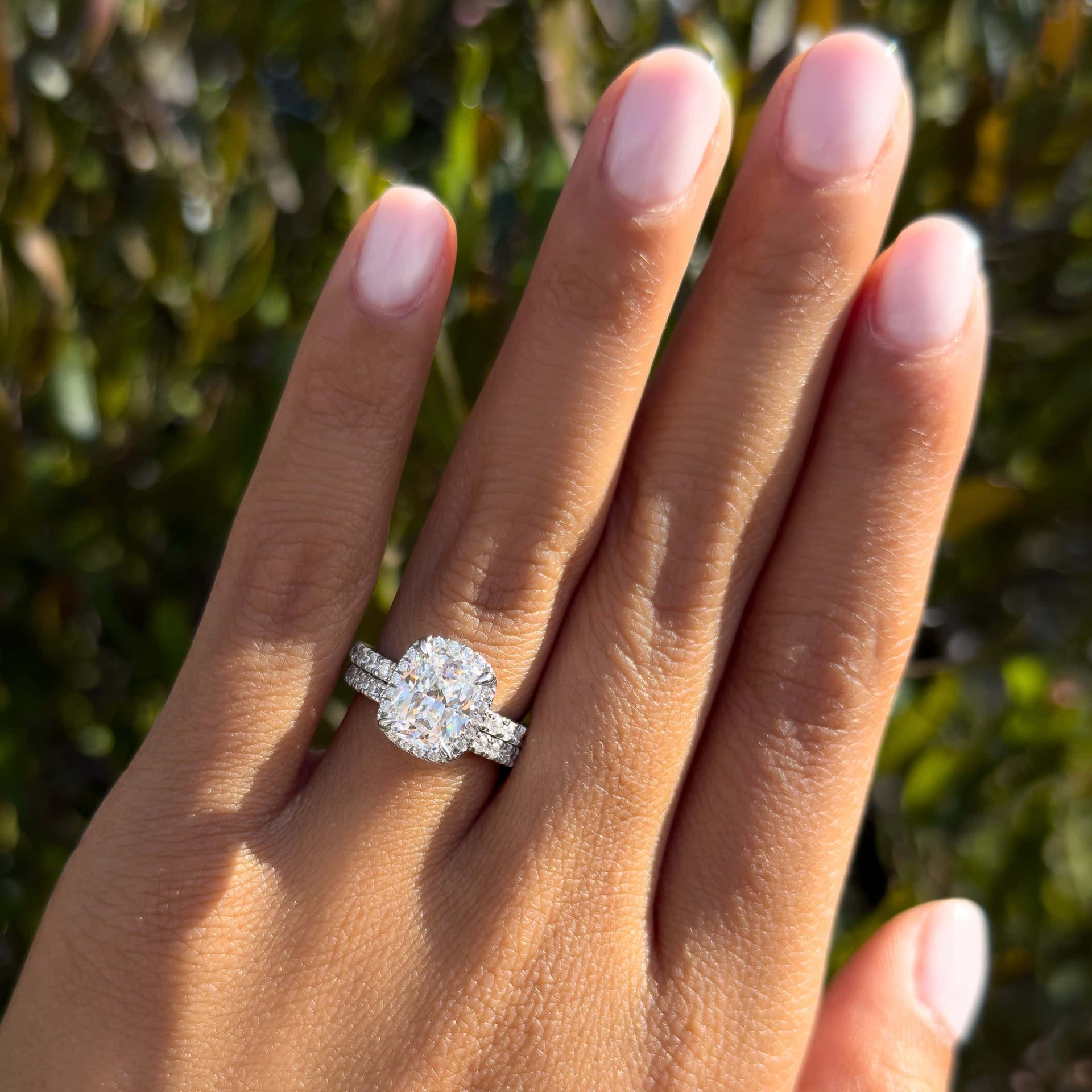 silver 3 carat elongated cushion cut halo engagement ring paired with matching wedding band worn by model in front of greenery