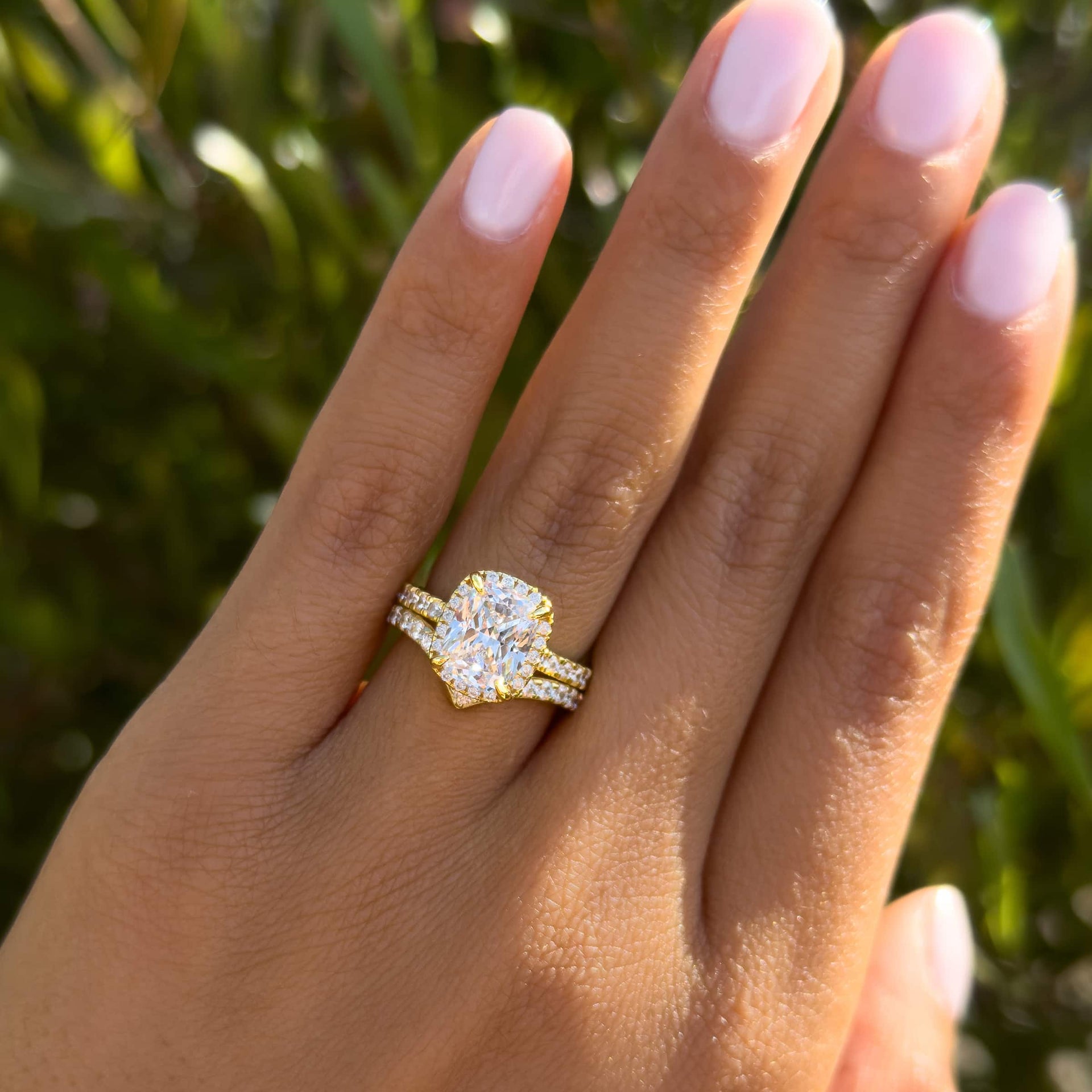 gold 3 carat elongated cushion cut halo engagement ring paired with chevron wedding band worn by model in front of greenery