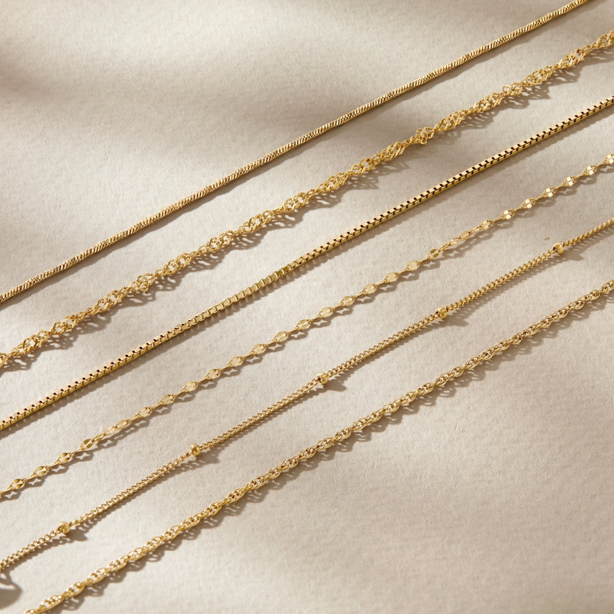 five 14k gold PVD plated stainless steel dainty chain bracelets resting across a tan surface 