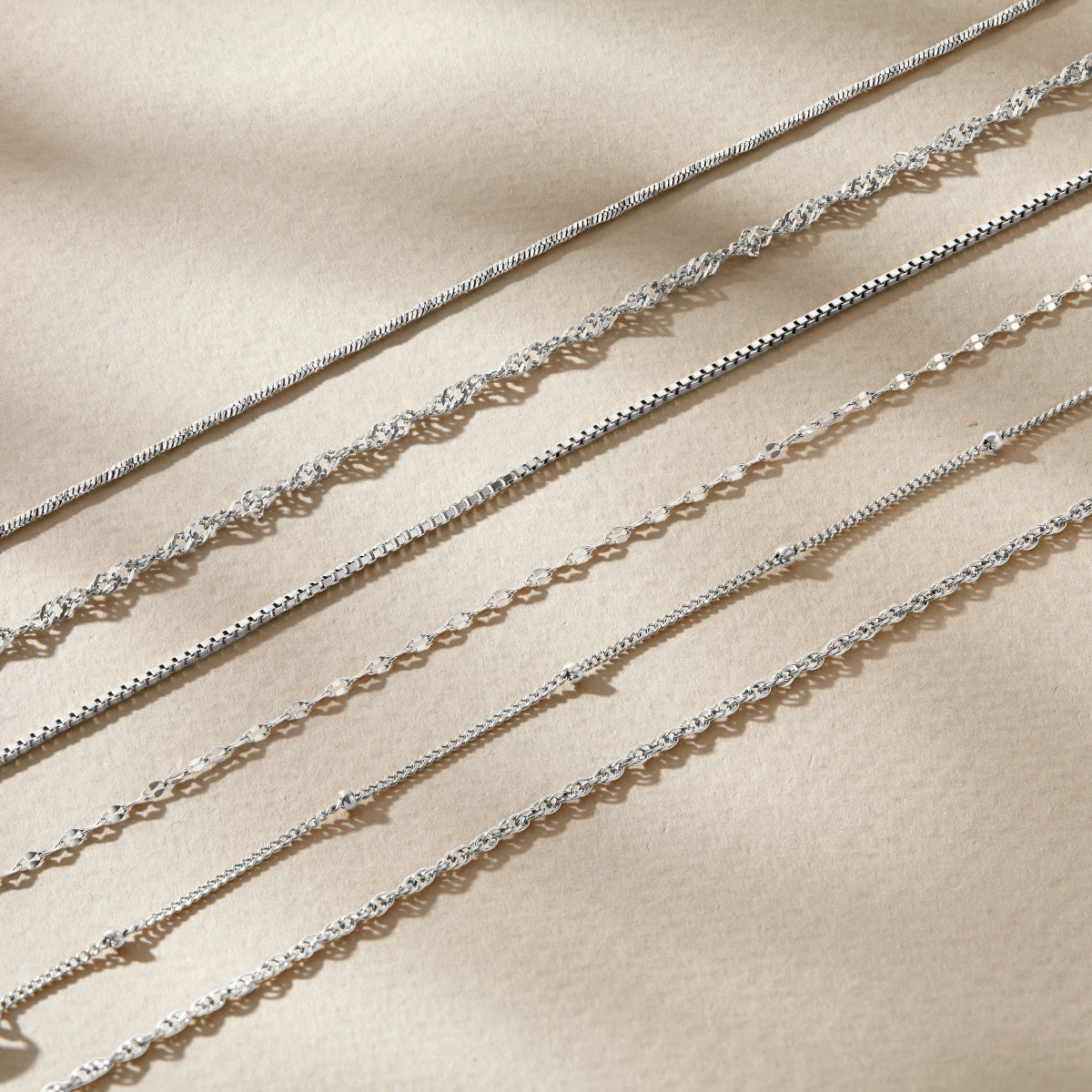 five silver stainless steel dainty chain bracelets resting across a tan surface 