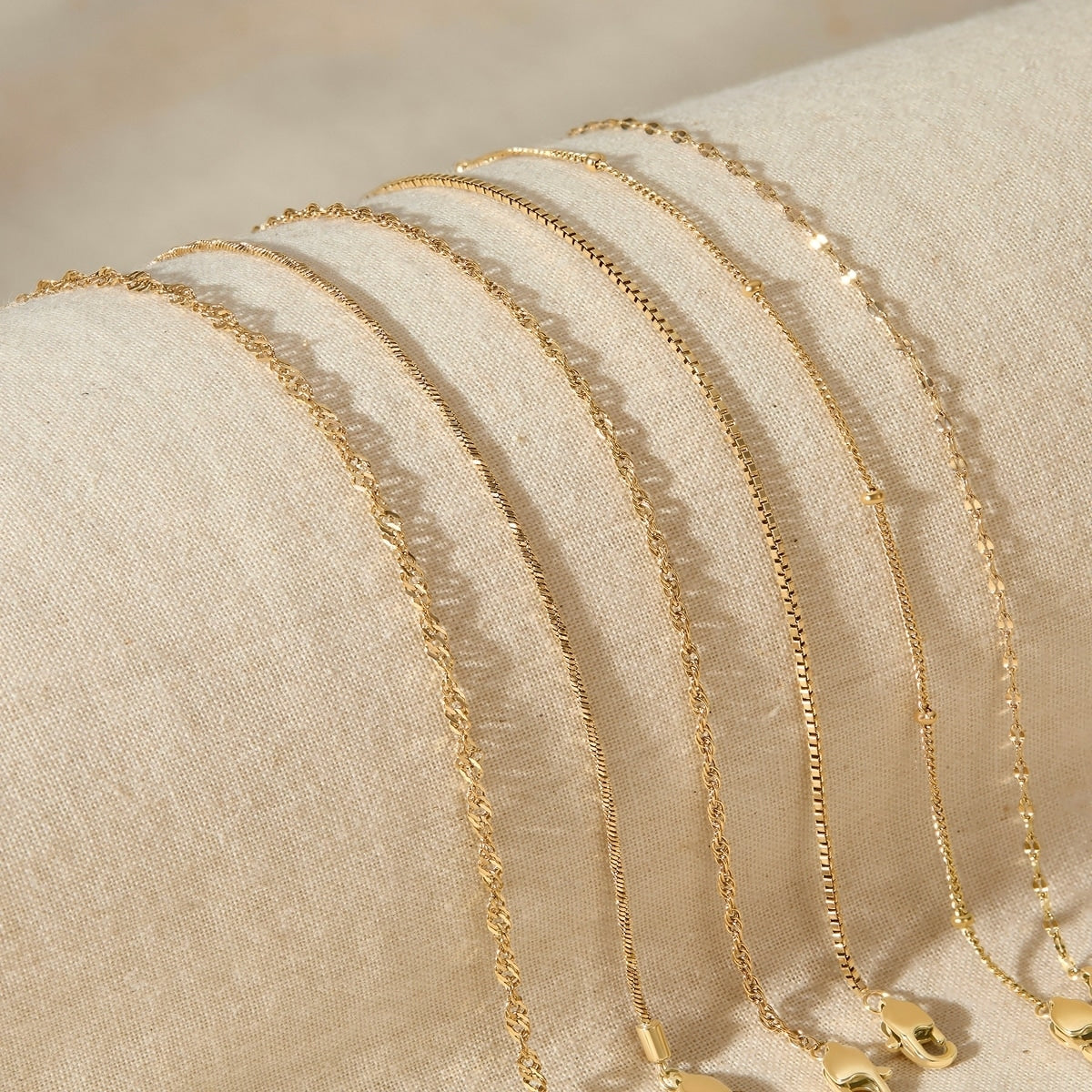 five gold stainless steel dainty chain bracelets resting across a tan surface 