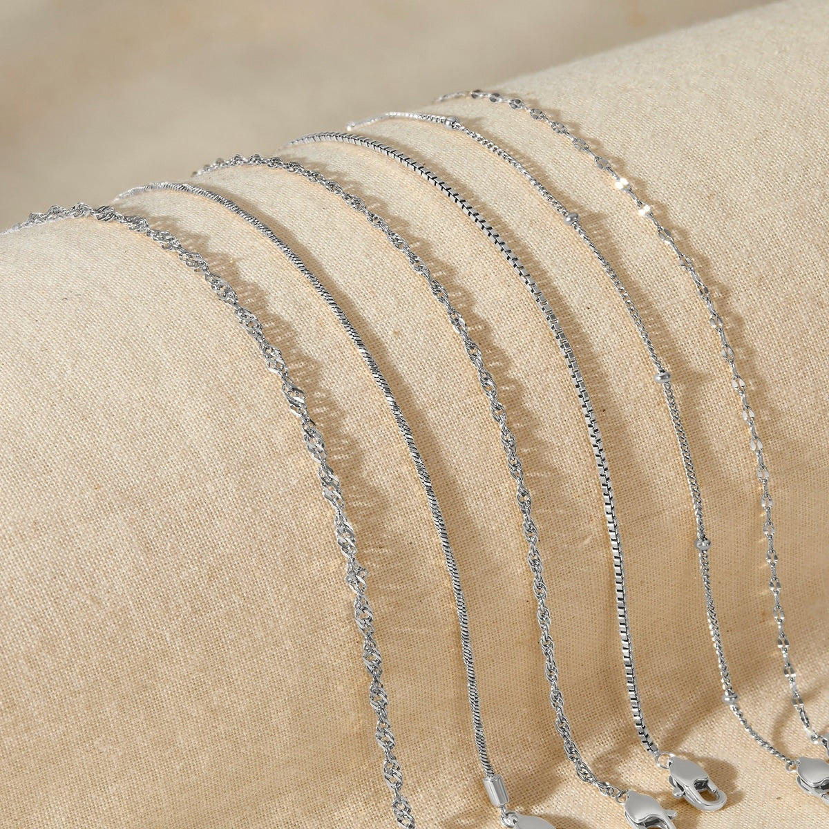 five silver stainless steel dainty chain bracelets resting across a tan surface 