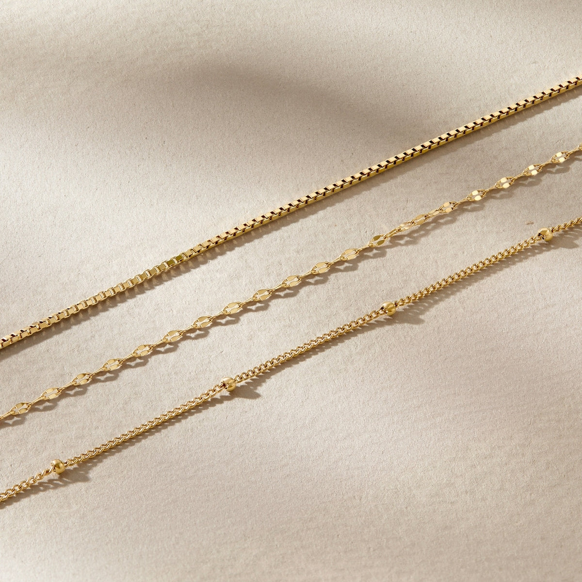 a flatlay of a 14k gold pvd plated stainless steel satellite chain, diamond cut lip chain, and box chain on a neutral, textured background