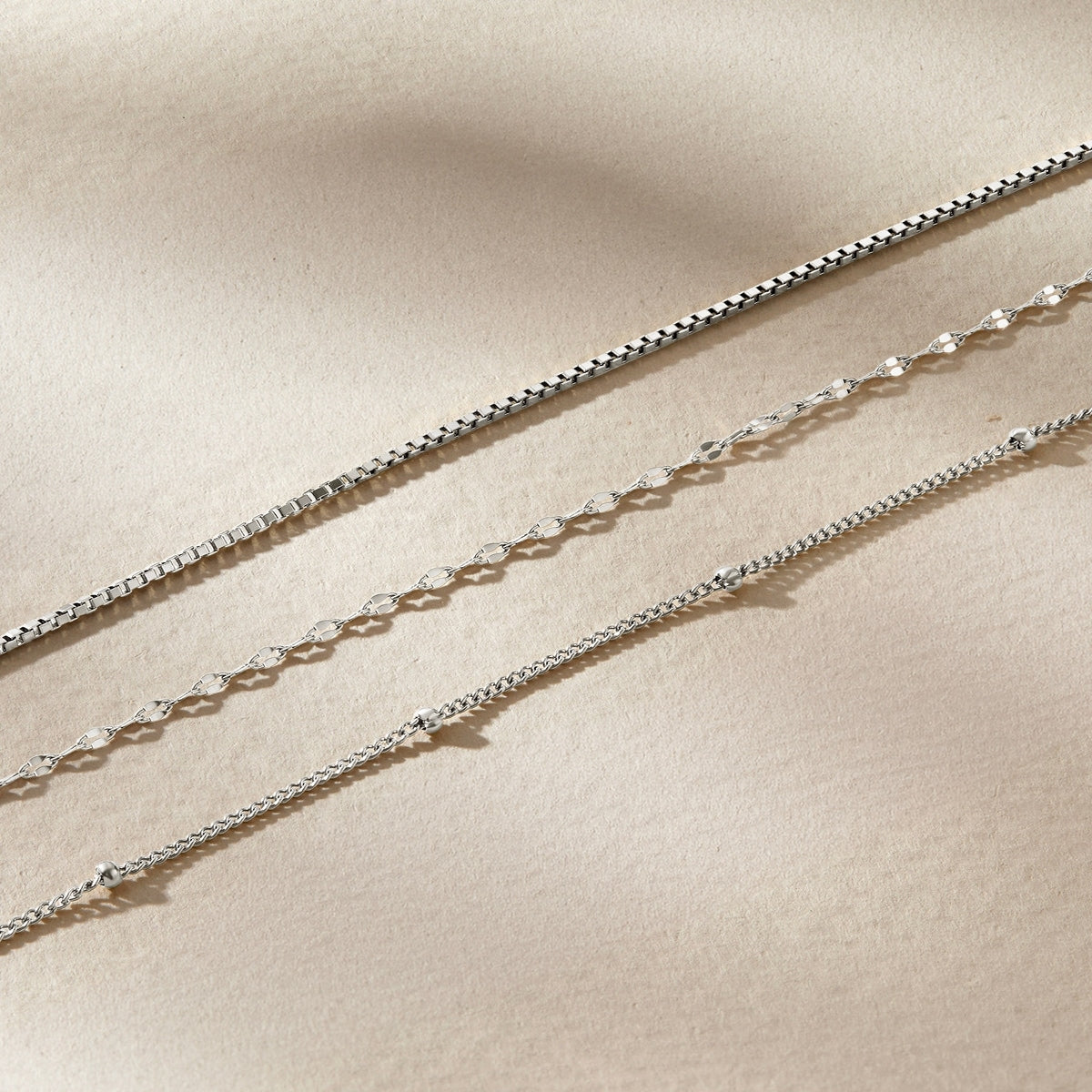 a flatlay of a stainless steel satellite chain, diamond cut lip chain, and box chain on a neutral, textured background 