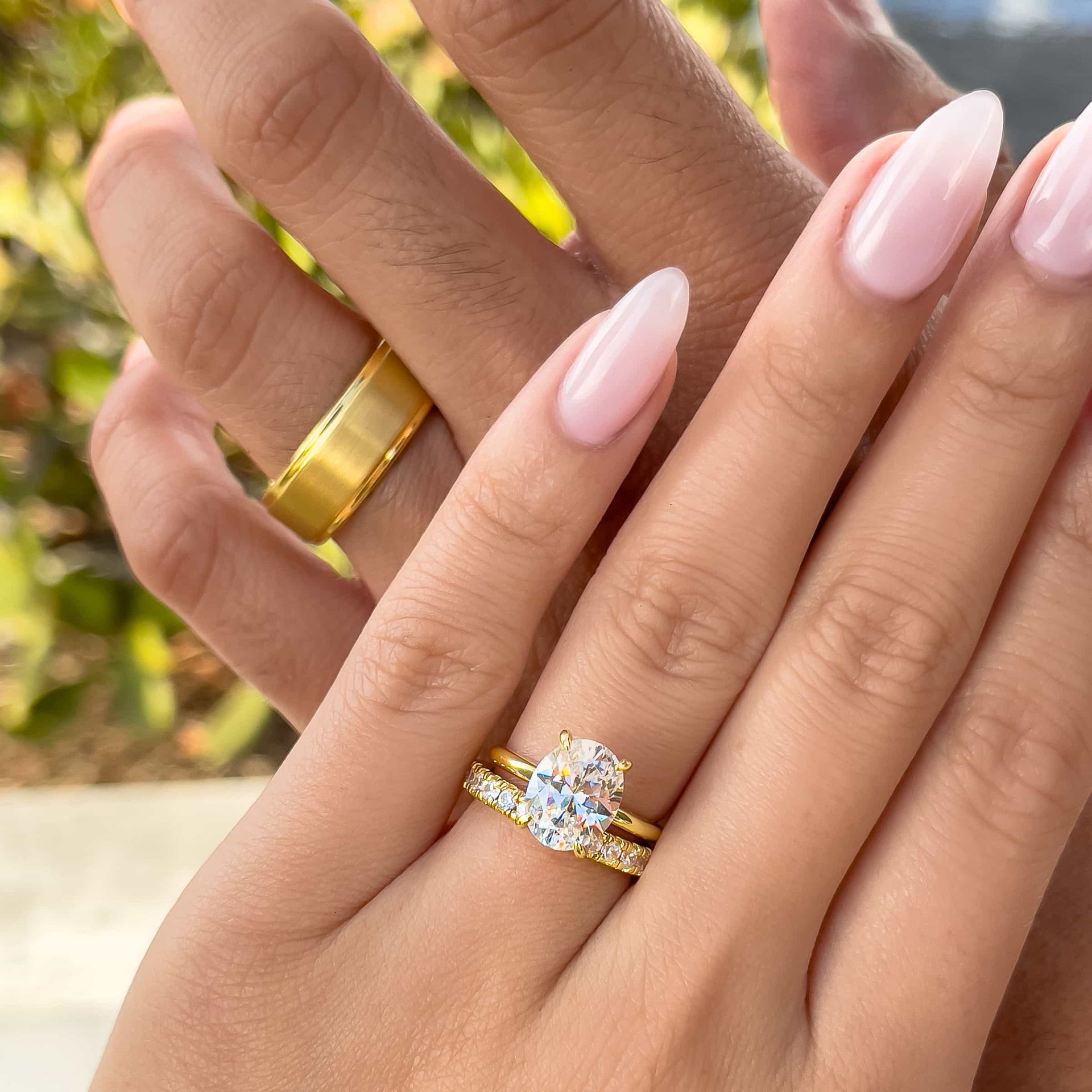 A his and hers gold wedding ring set, made up of a gold tungsten carbide men's wedding band and a gold oval cut solitaire ring paired with an eternity wedding band. Shown on male and female model.