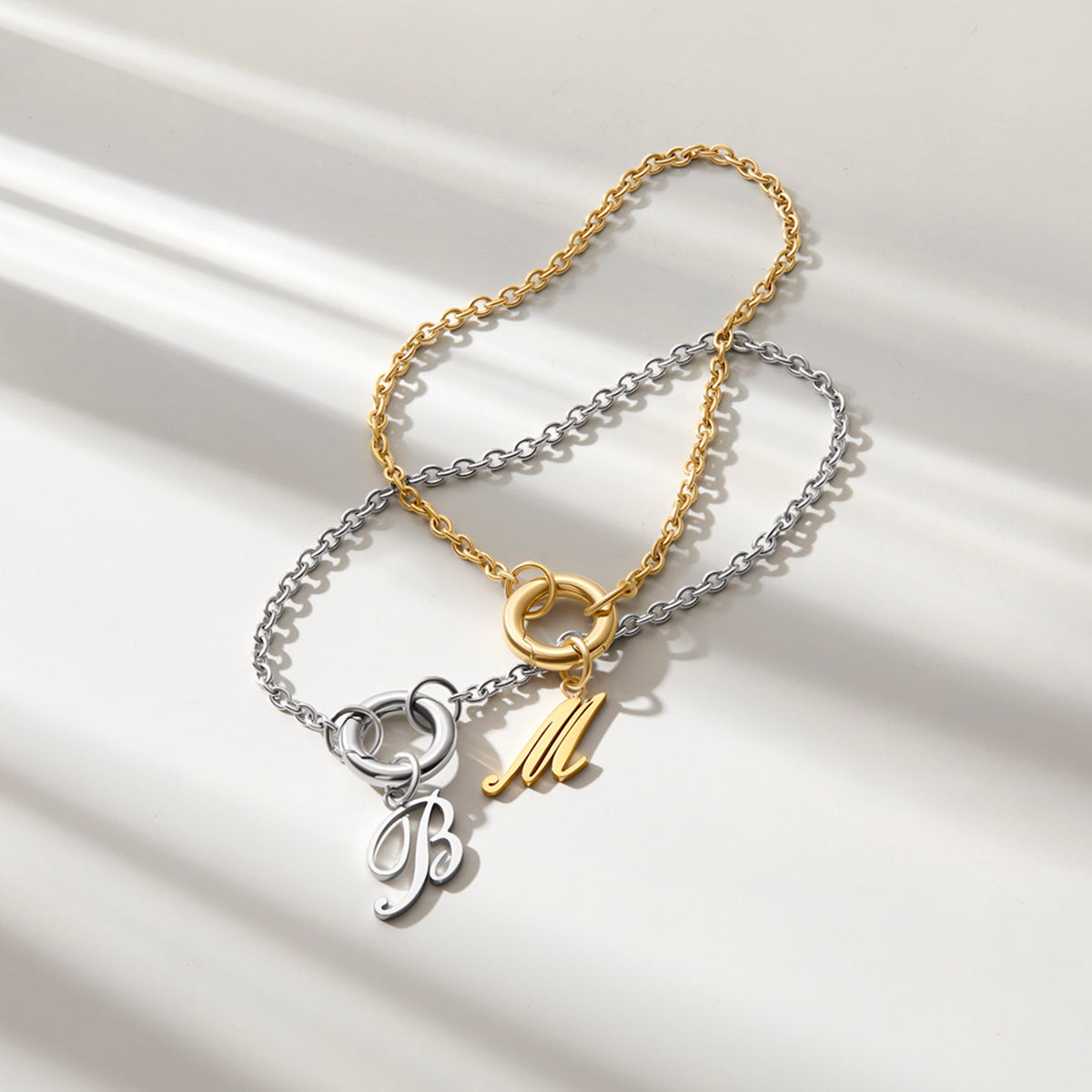 stainless steel cable chain charm bracelets shown in silver with a "B" initial charm & gold with an "M" initial charm posed overlapped on an off white background. 