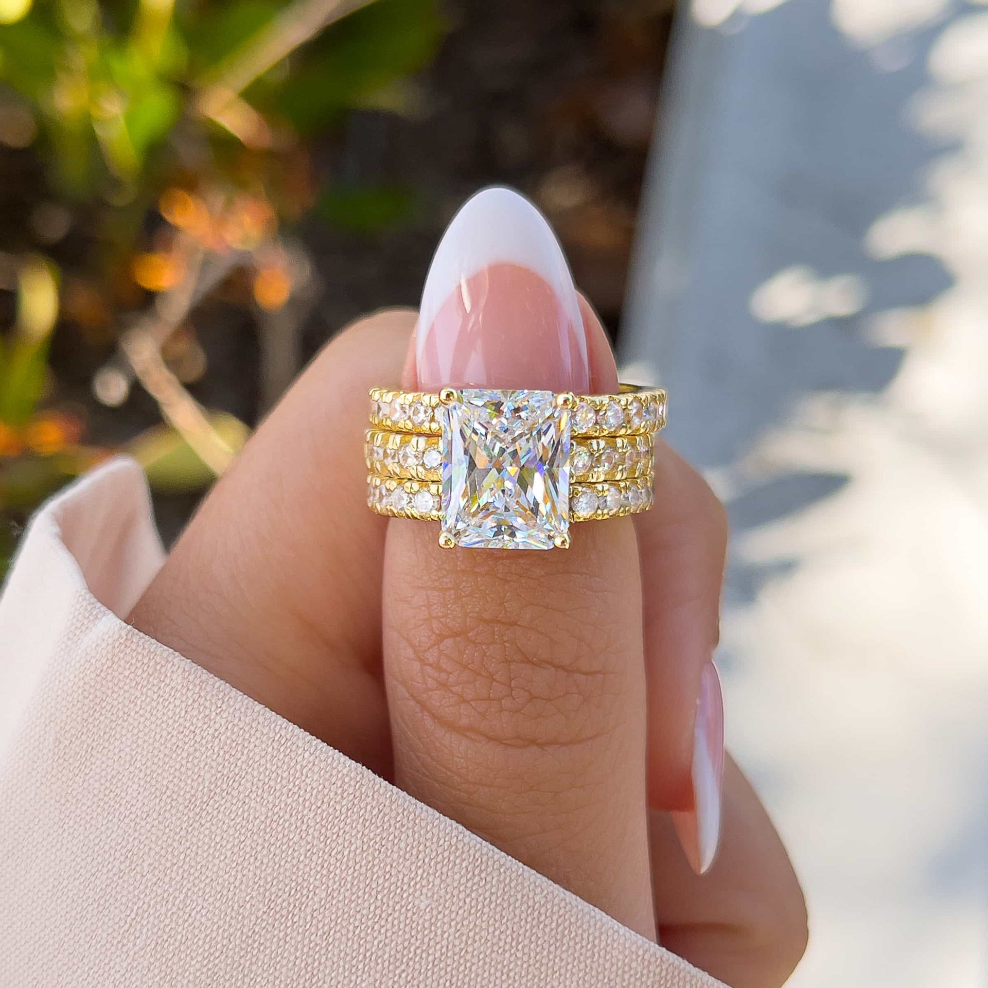 gold radiant triple wedding ring stack modeled on female hand with french tip nail and neutral sleeve with greenery in the background