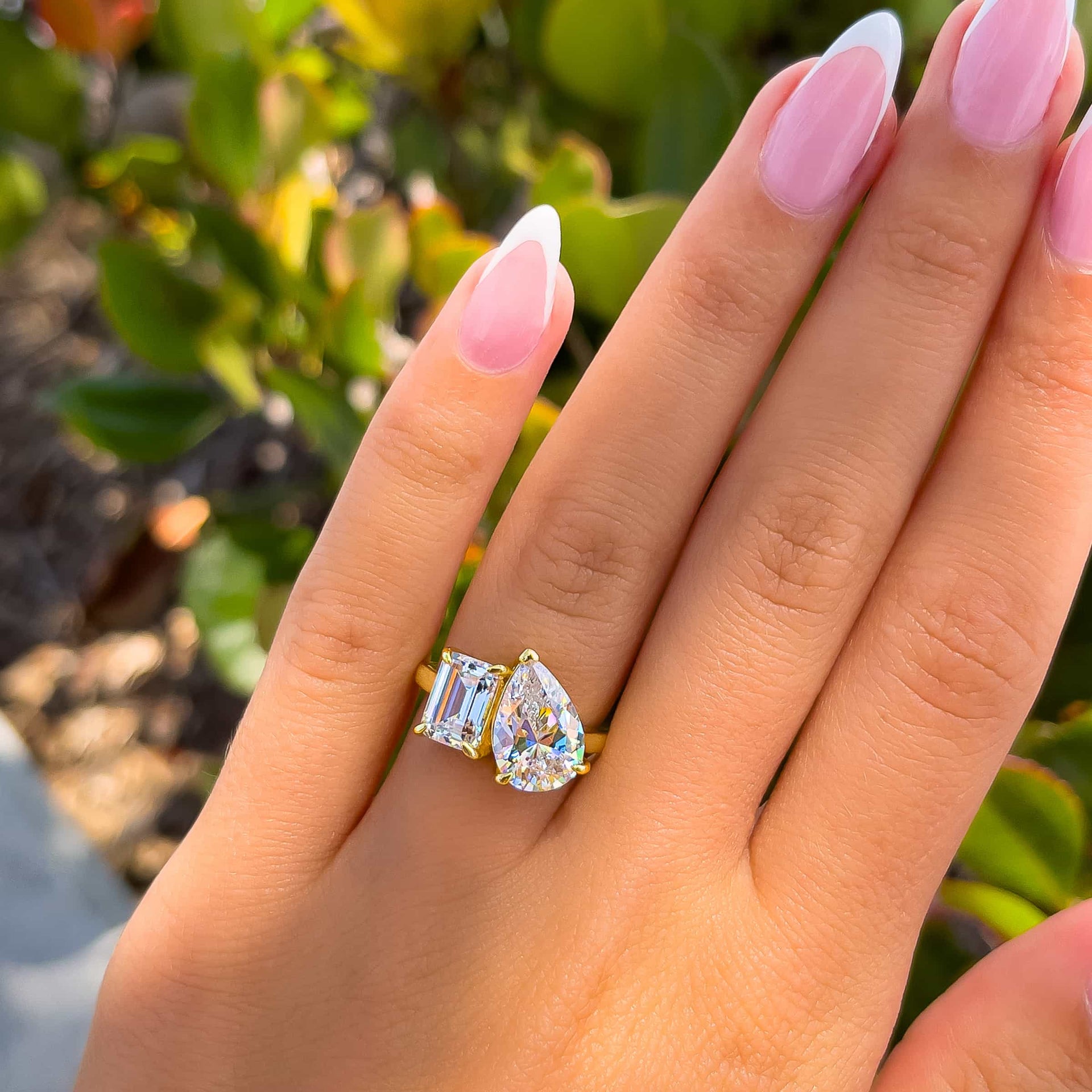 1.75 emerald cut and 4 carat pear cut two stone gold engagement ring on model with french tip nails