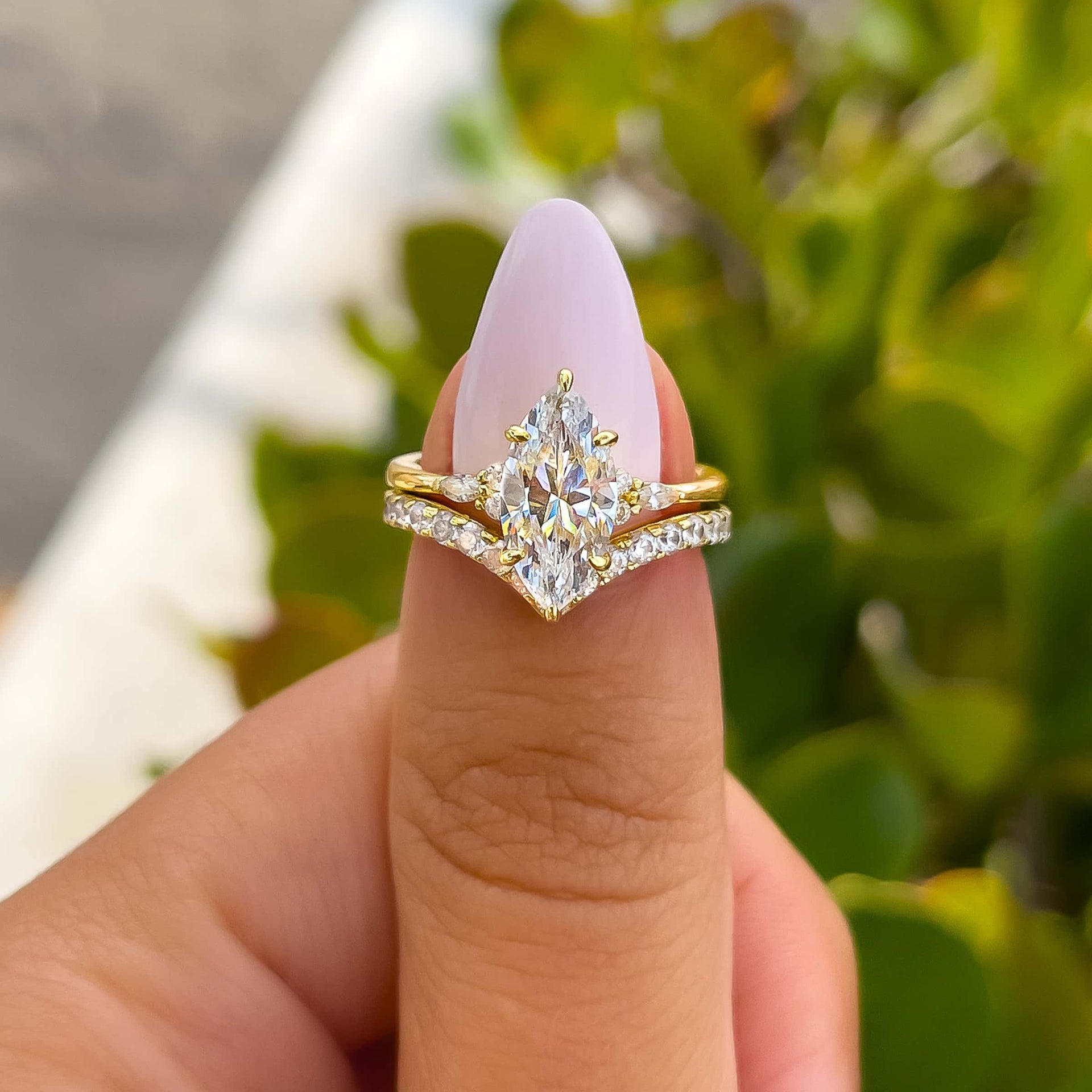 woman pinching marquise engagement ring with complimentary gold wedding band outside with greenery shown in the background