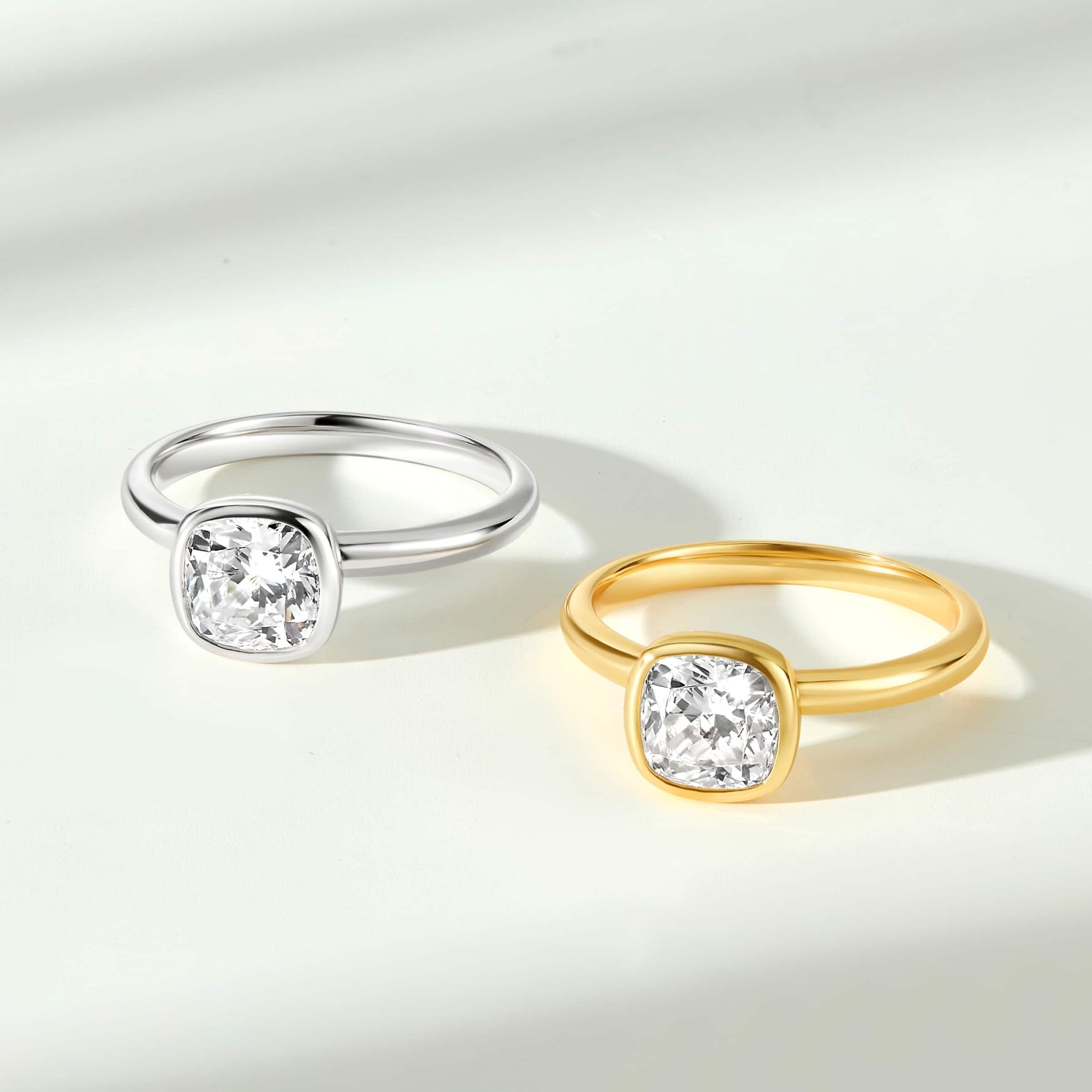 flat lay image of two cushion cut engagement rings with bezel settings sitting side-by-side on a white background