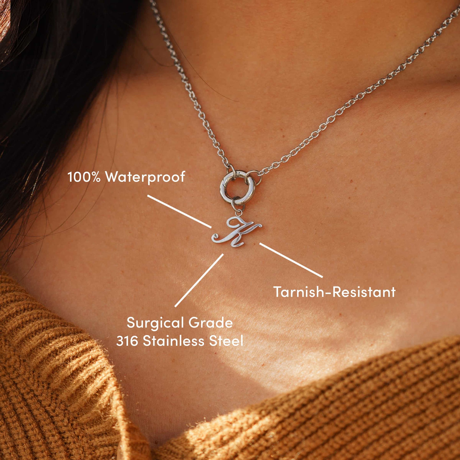 minimalist silver charm necklace with an "K" cursive letter charm on a model wearing a brown sweater with the following text: "Tarnish-Resistant", "100% Waterproof", "Surgical Grade 316 Stainless Steel"