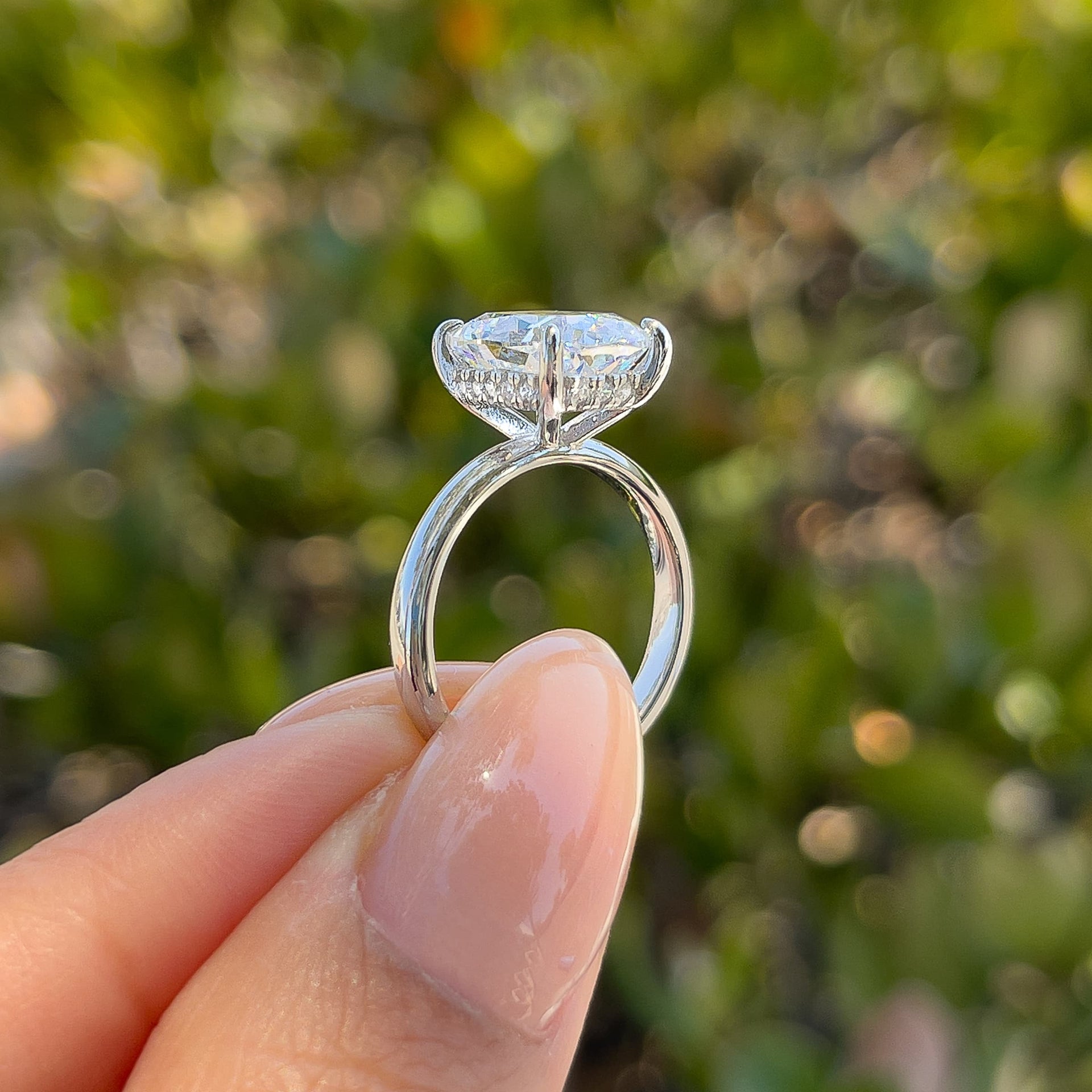 side view of silver 4 carat elongated cushion cut solitaire engagement ring with hidden halo held by model with neutral nails in front of greenery