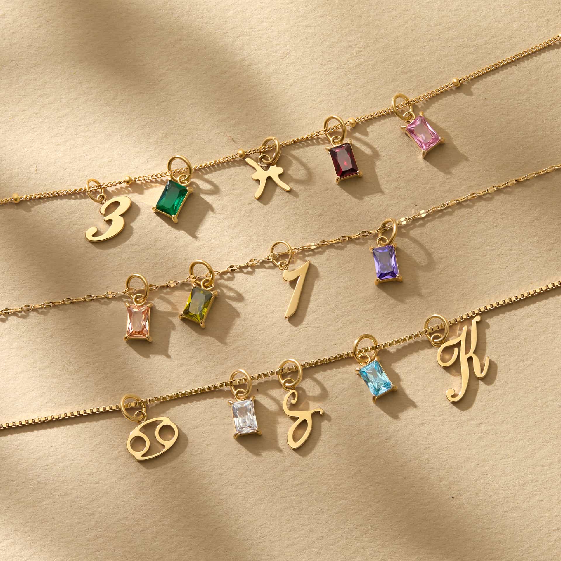 a stunning flatlay of a variety of 14k Gold PVD plated stainless steel charms, all modeled on various chain necklaces atop a tan and textured background