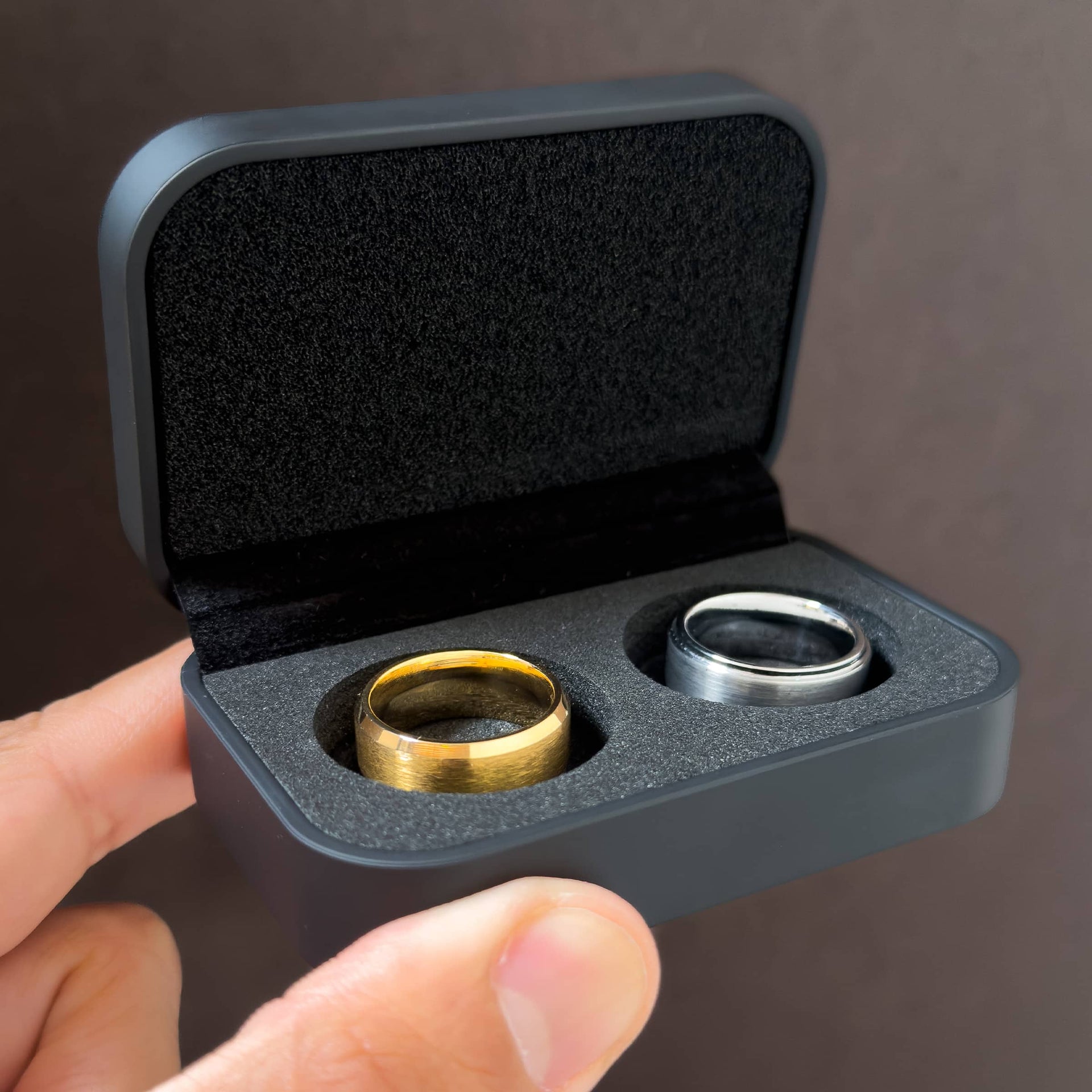 a sleek black ring box held by a model in front of a dark background, opened to show a black padded interior and gold and silver men's bands inserted in the slots