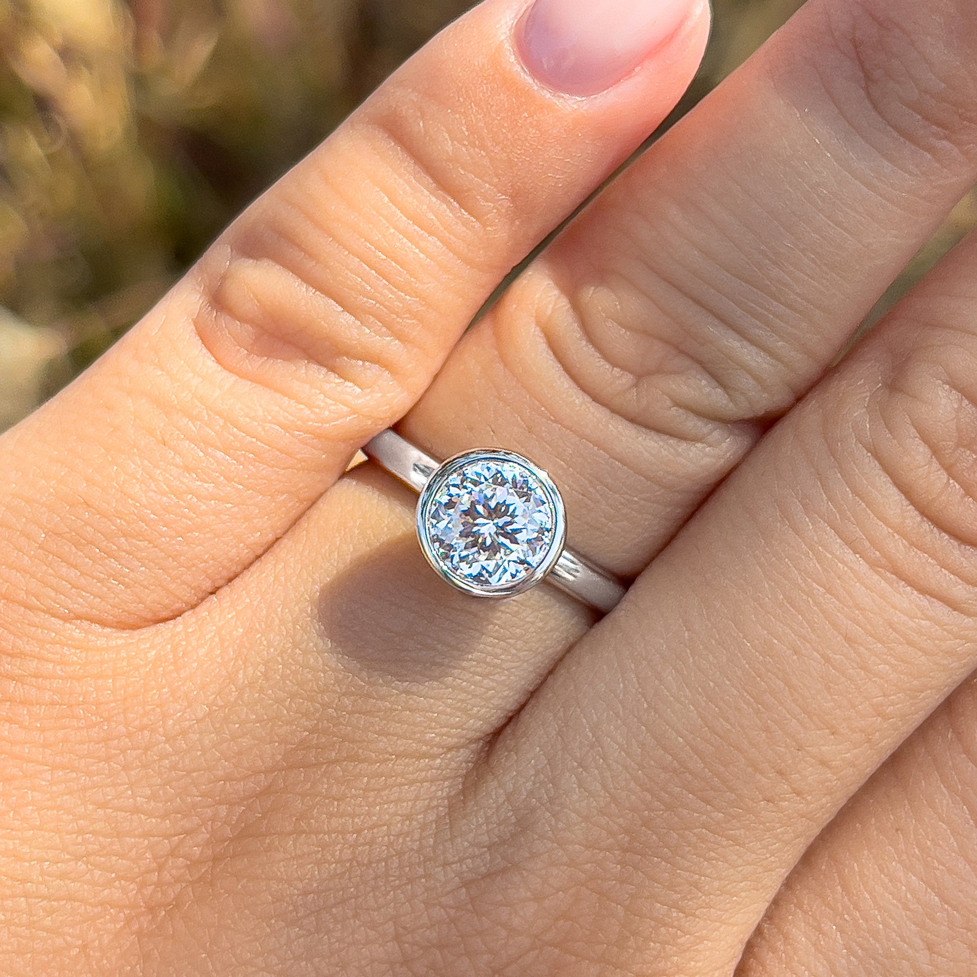 a close-up shot of a silver bezel round cut ring on model's ring finger with pink manicured nails
