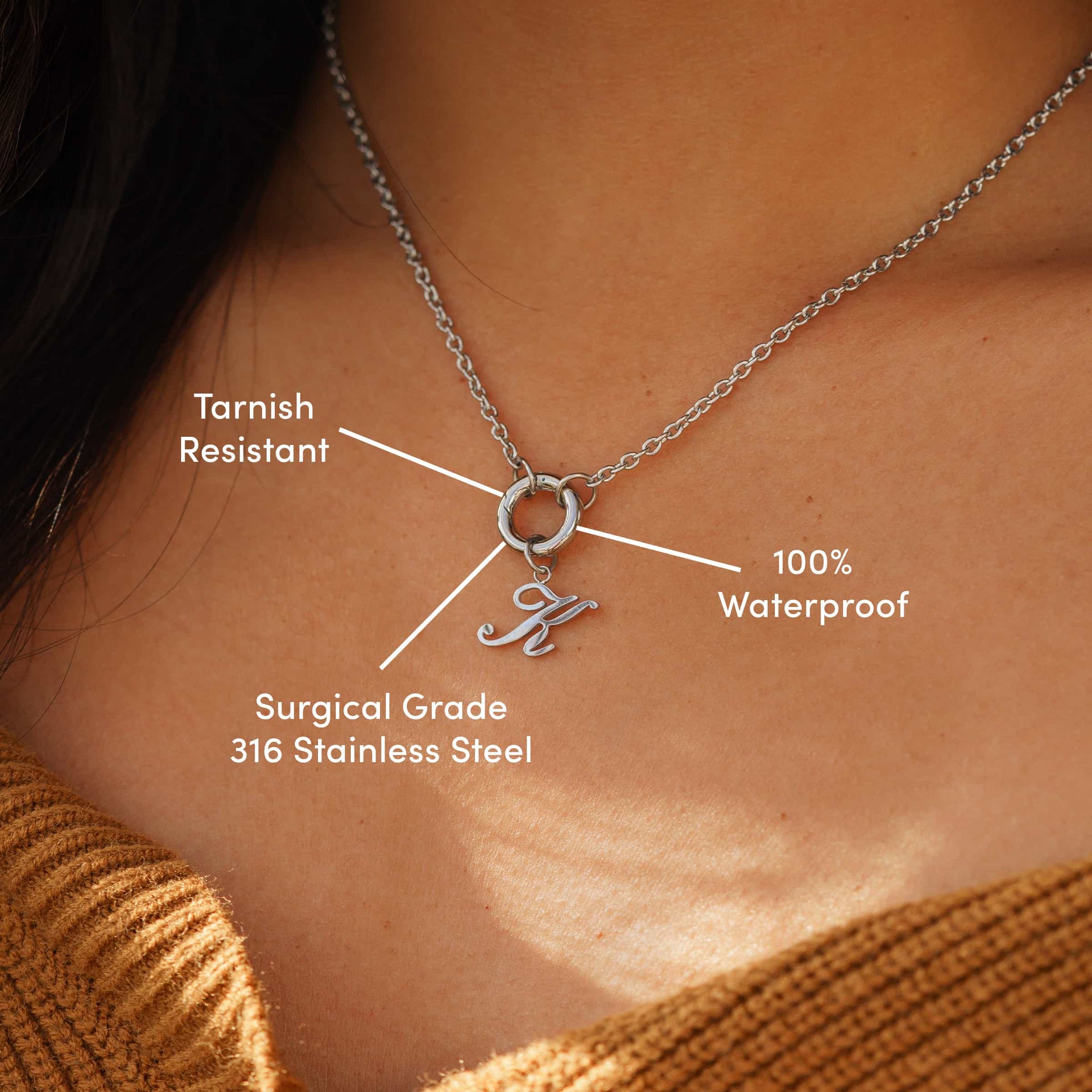 minimalist silver charm necklace with an "K" cursive letter charm on a model wearing a brown sweater with the following text: "Tarnish Resistant", "100% Waterproof", "Surgical Grade 316 Stainless Steel"