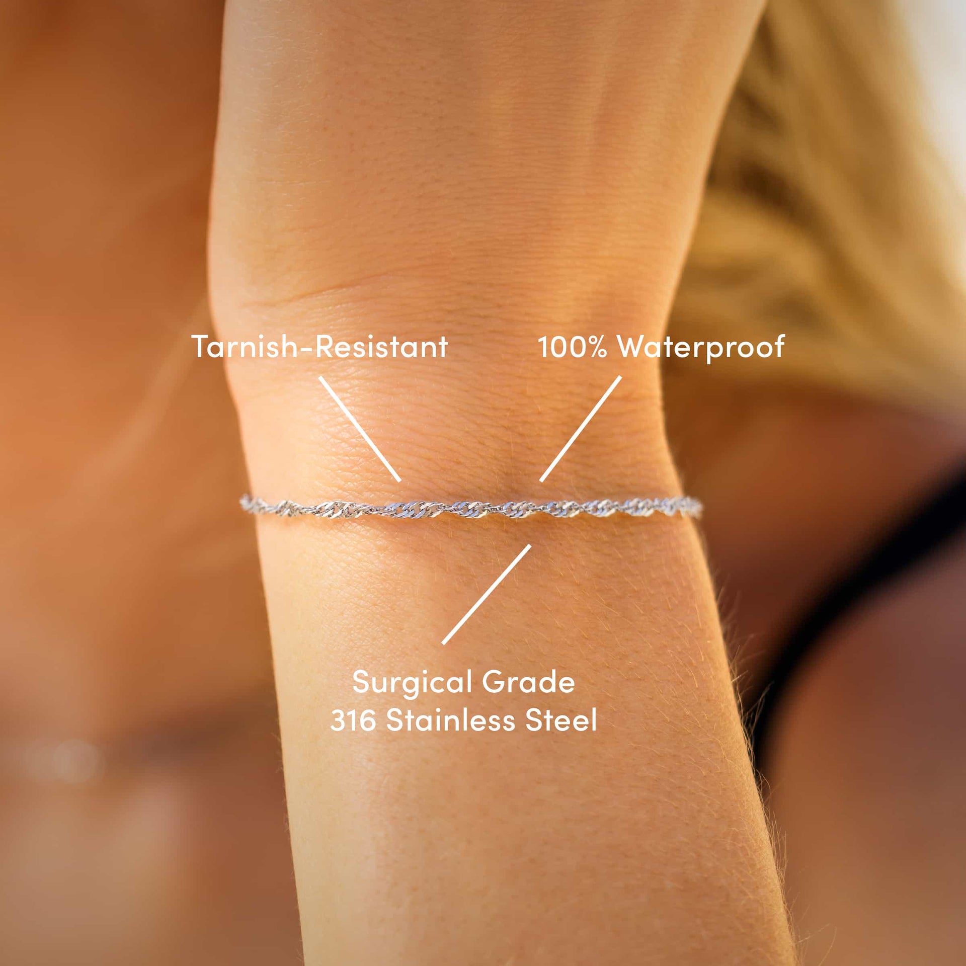 a close up of a dainty stainless steel Singapore chain bracelet worn by a model with the text overlayed "tarnish-resistant, 100% waterproof, & surgical grade 316 stainless steel" 