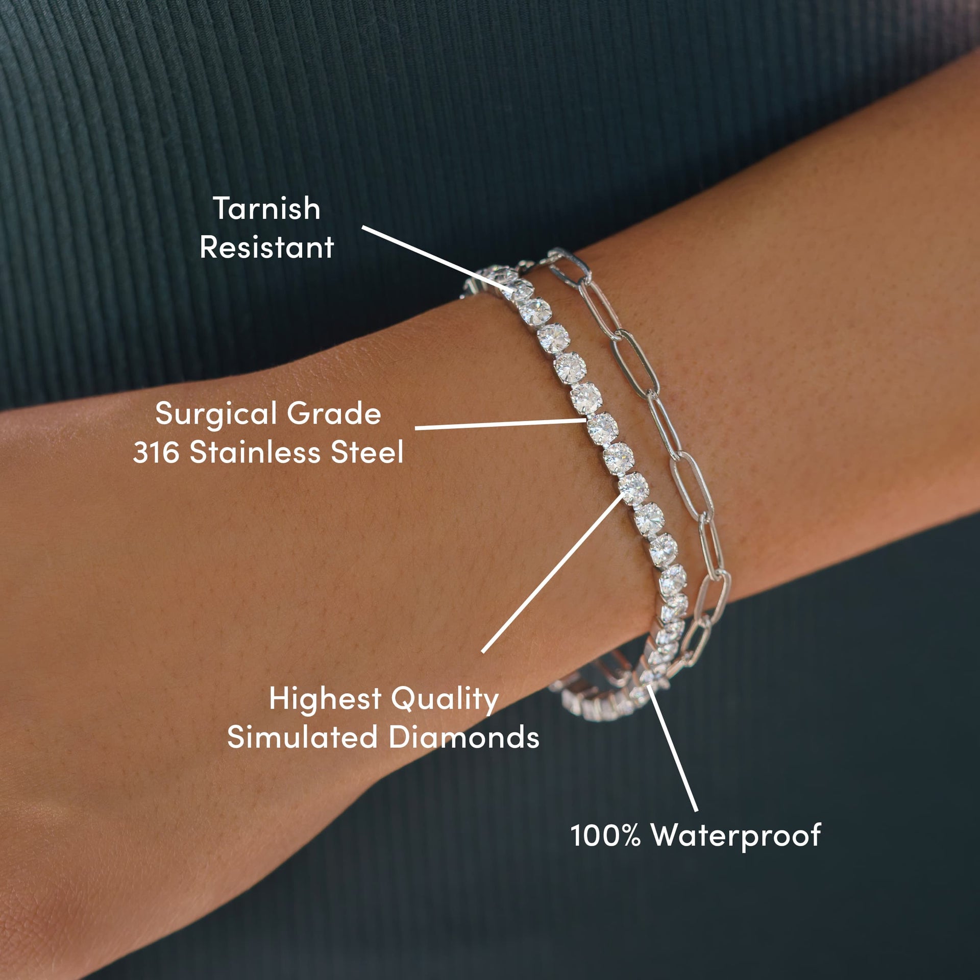 everyday silver tennis bracelet worn as a part of a bracelet stack with a silver paperclip necklace by model wearing dark colors with the text over it: Tarnish Resistant, Surgical Grade 316 Stainless Steel, Highest Quality Simulated Diamonds, 100% Waterproof