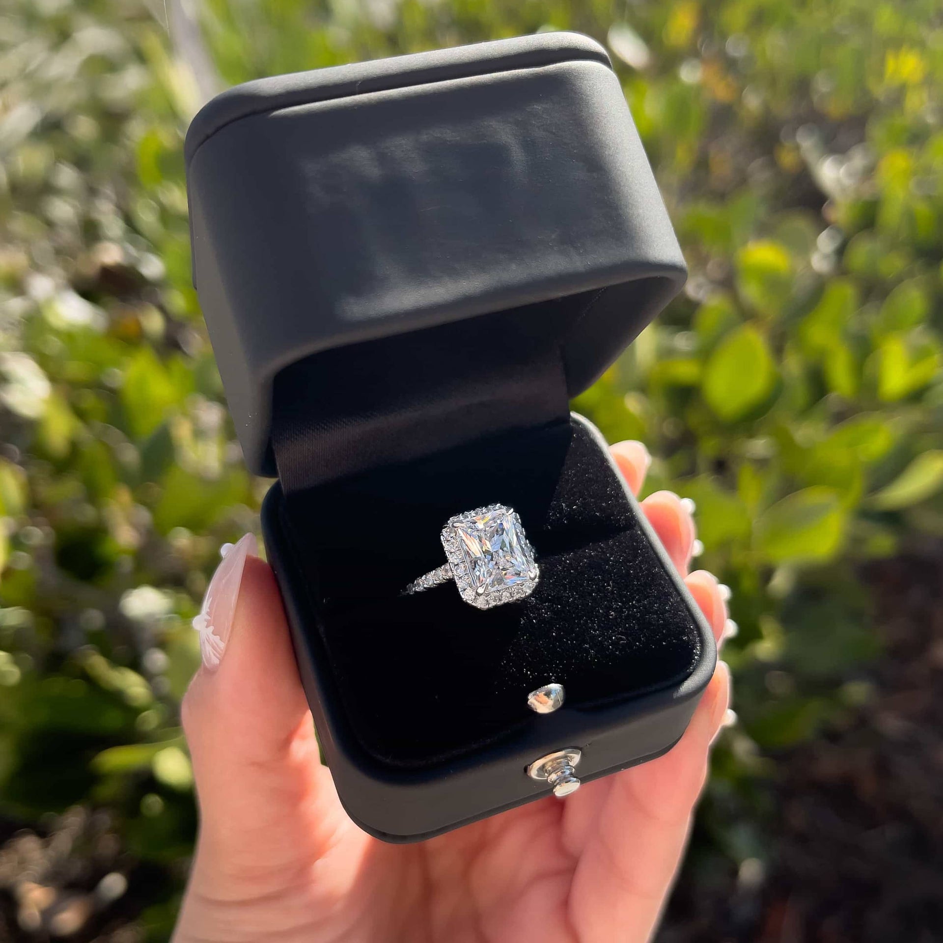 simple matte black ring box in a front-facing, open position with a stunning silver radiant cut engagement ring inside held by model in front of greenery