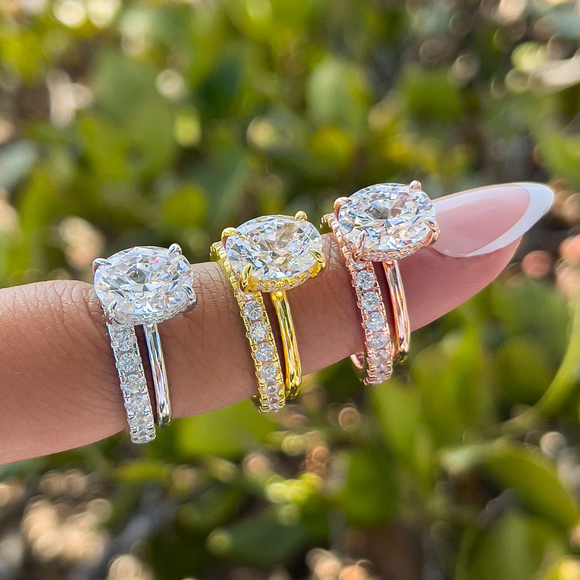 a classic 3 carat oval cut wedding set shown in silver, gold, and rose gold on model with french tip nails in front of greenery
