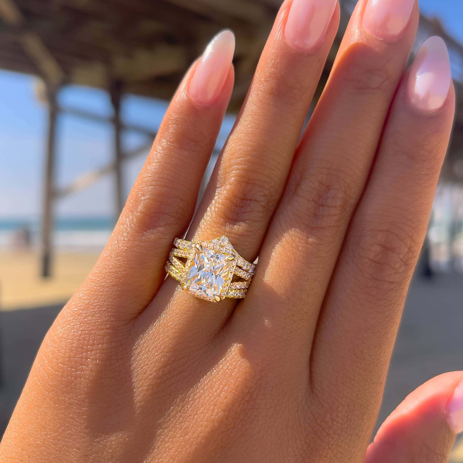 3.5ct radiant cut engagement ring with two complimentary wedding bands on females hand by ocean