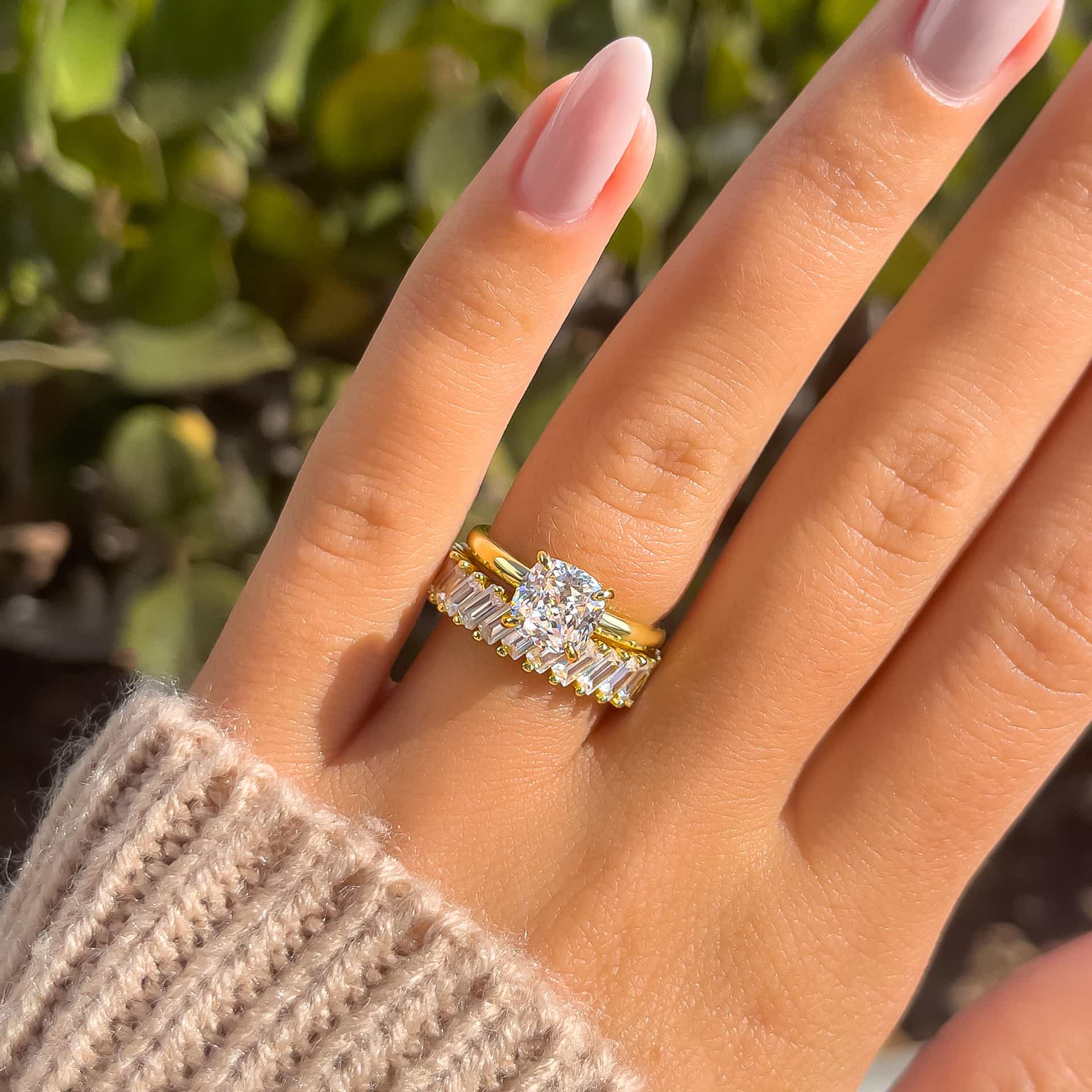 a shiny gold wedding ring stack made up of a 1.5 carat cushion cut solitaire and a baguette wedding band on model with light pink nails and tan sweater