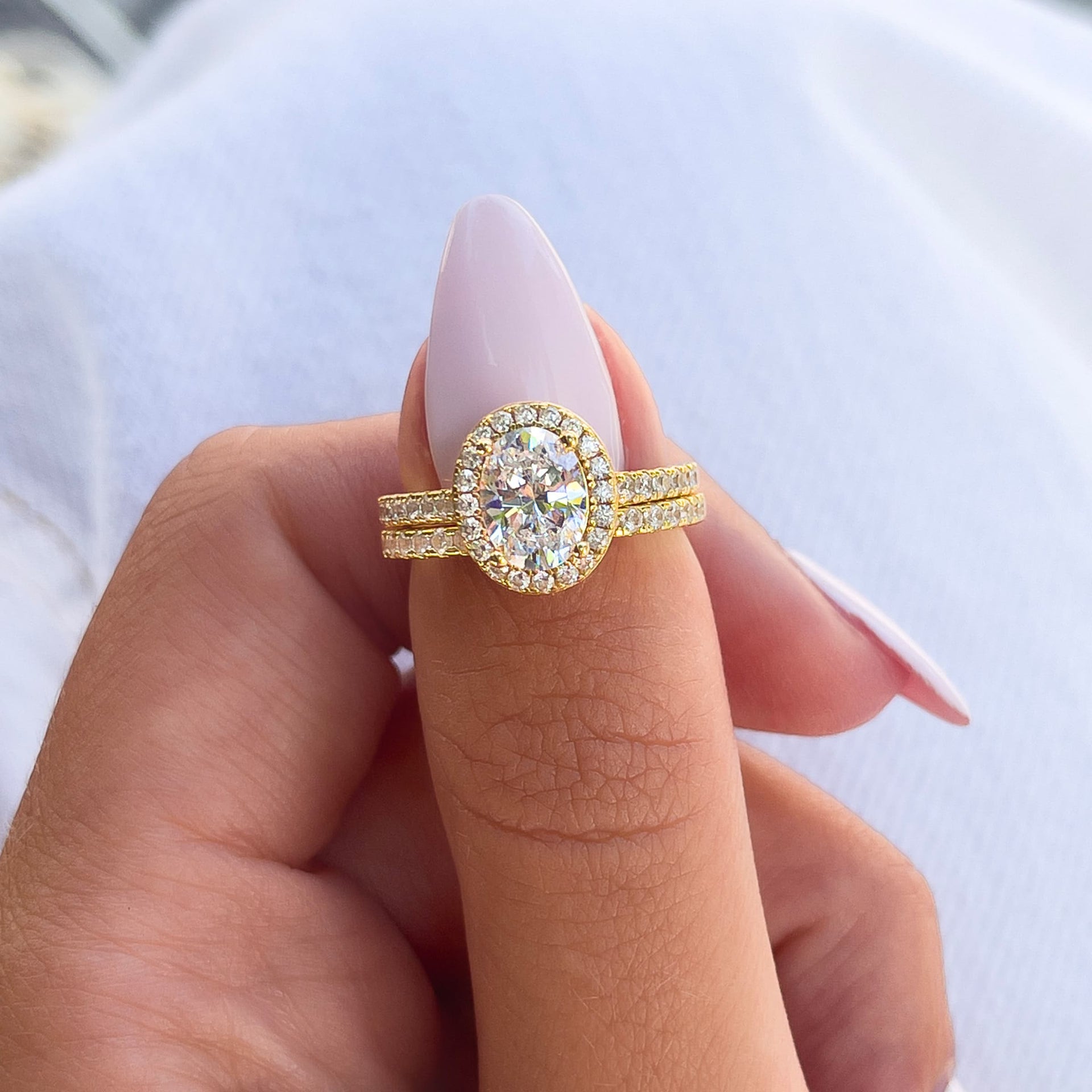 gold oval cut engagement ring with halo and matching gold wedding band on female model with light pink nails