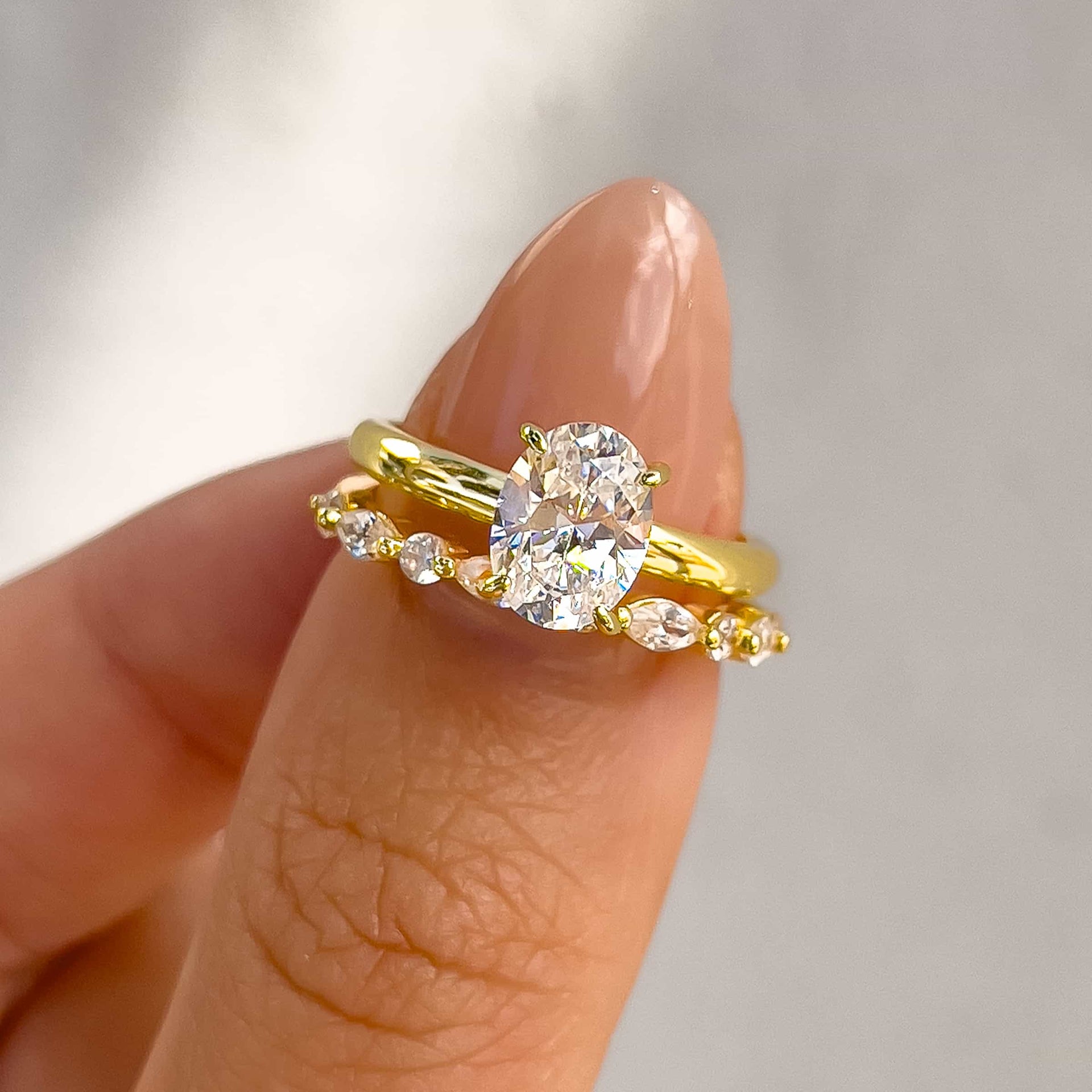 gold pave wedding band with round and marquise stones paired with oval solitaire engagement ring held by model with pink nails on gray background