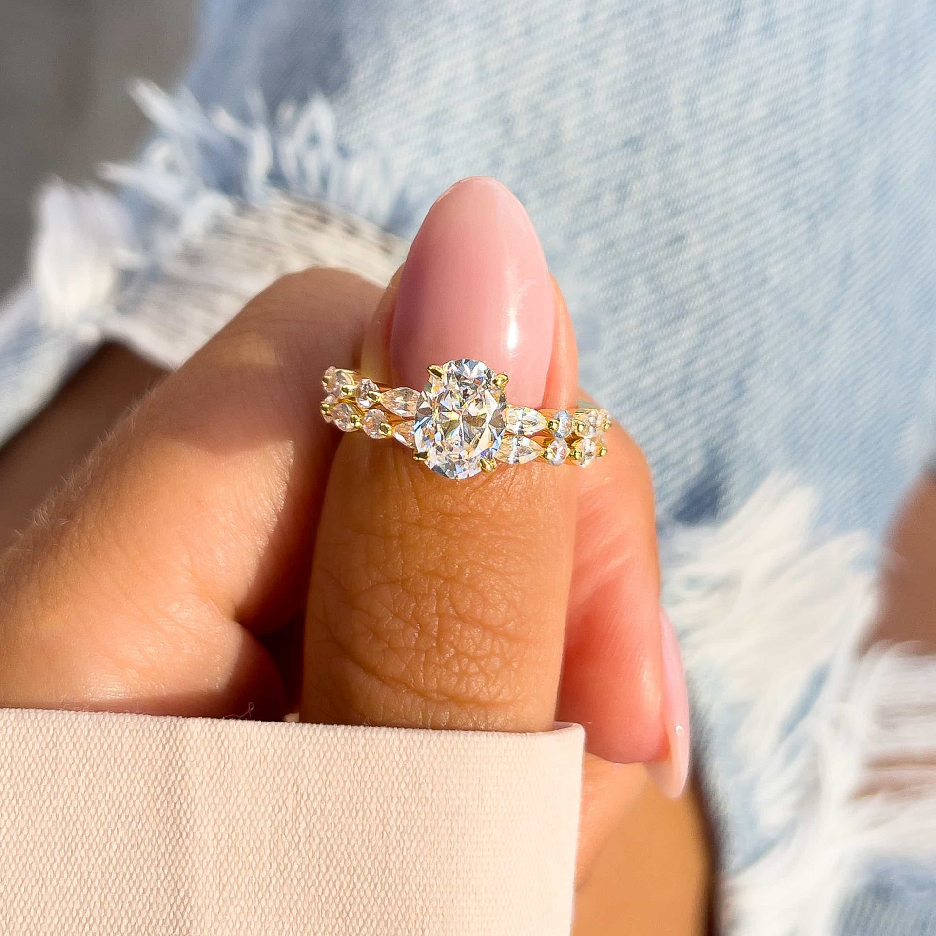 gold oval engagement ring with gold pave wedding band on model with pink nails and tan sleeve with blue jeans in the background