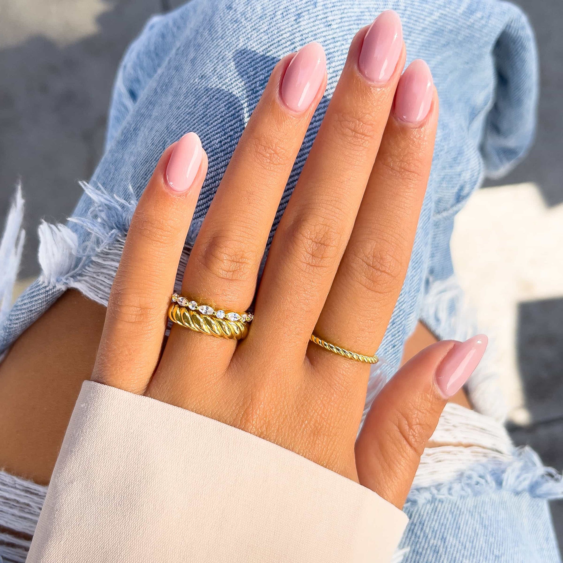 three gold wedding bands on models hand with pink nails with blue jeans and tan sleeve