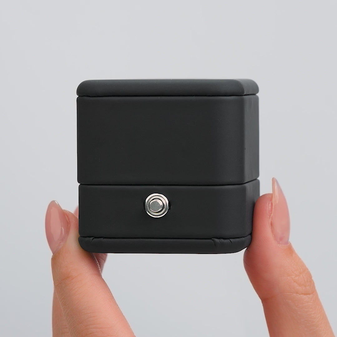 video of a matte black ring box with a black interior and silver push-to-open button with a silver engagement ring that gets placed inside of it by model