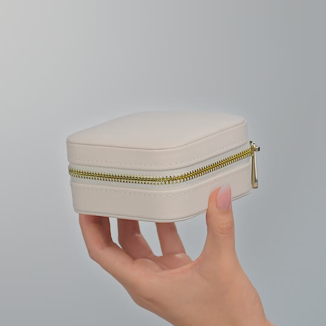 A video of a square shaped travel jewelry box with gold hardware shown in its cream and charcoal colored variants in various positions. Held by a model against an off white background
