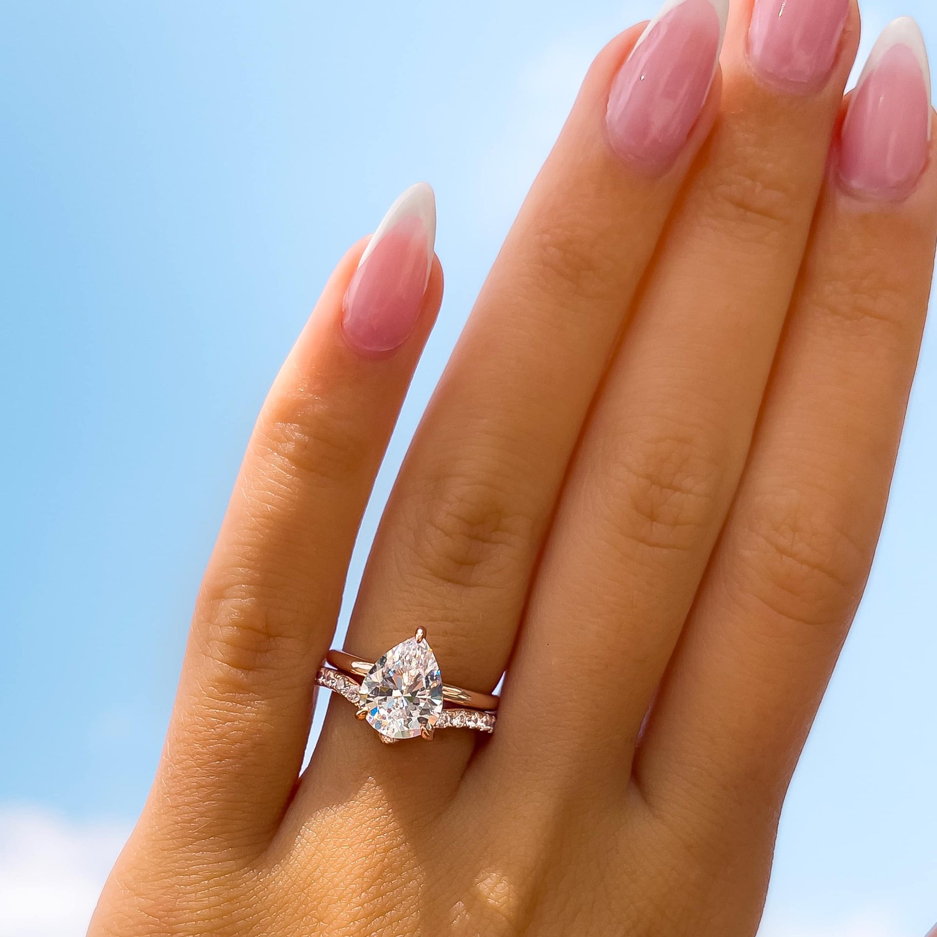 3 carat rose gold pear cut engagement ring with a hidden halo paired with a v-shaped half eternity wedding band on a female hand that is up towards the sky