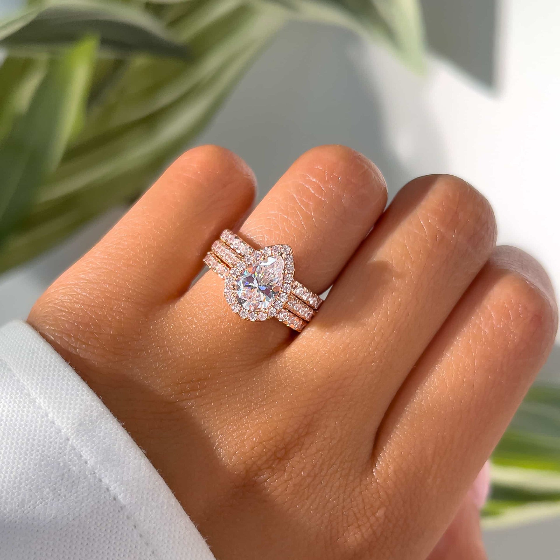 2 carat rose gold pear cut engagement ring triple stack on female hand in front of greenery and a white wall