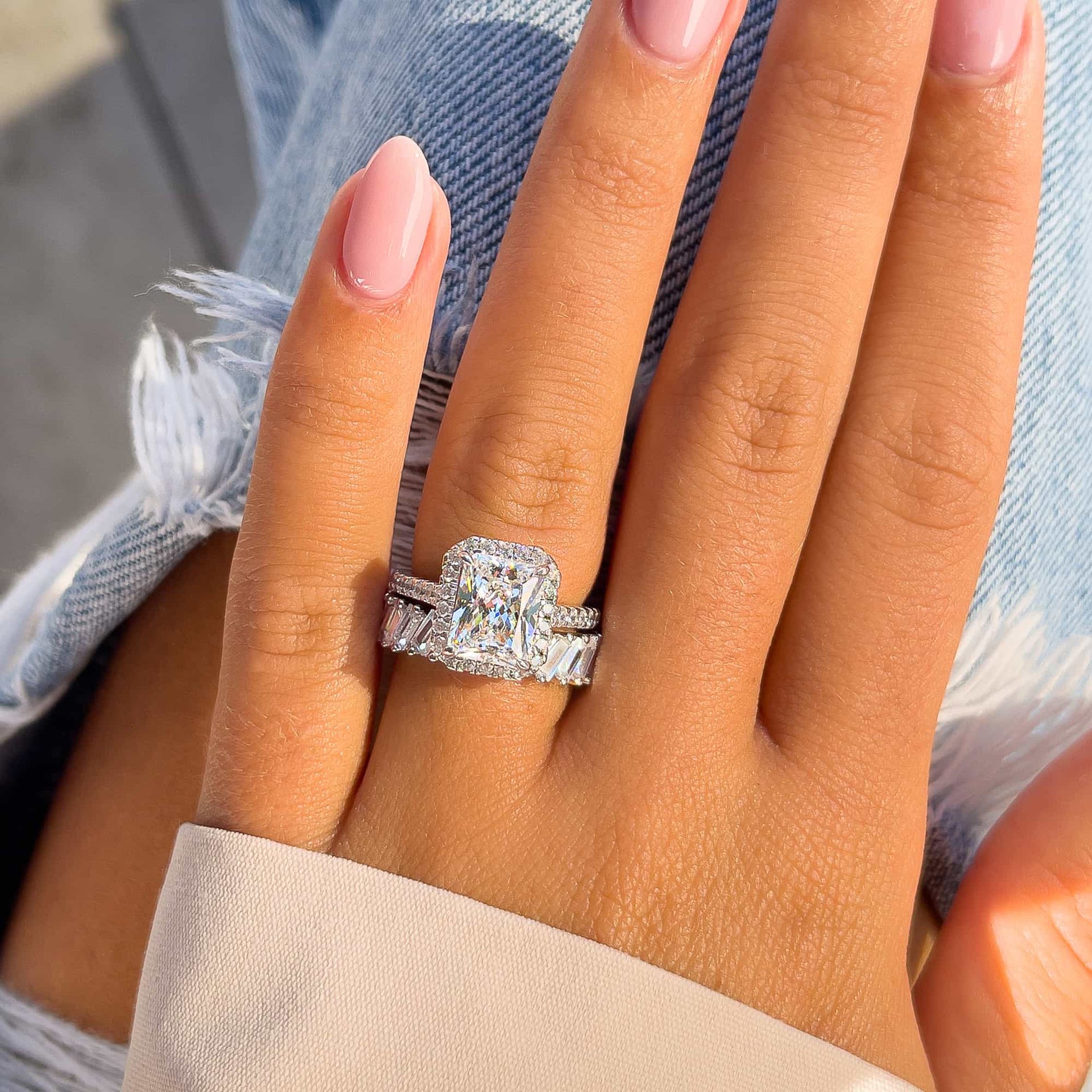 stunning 3.5 carat silver radiant cut engagement ring paired with a  baguette band with inset diamonds on model's hand with pink nails and a cream colored sleeve