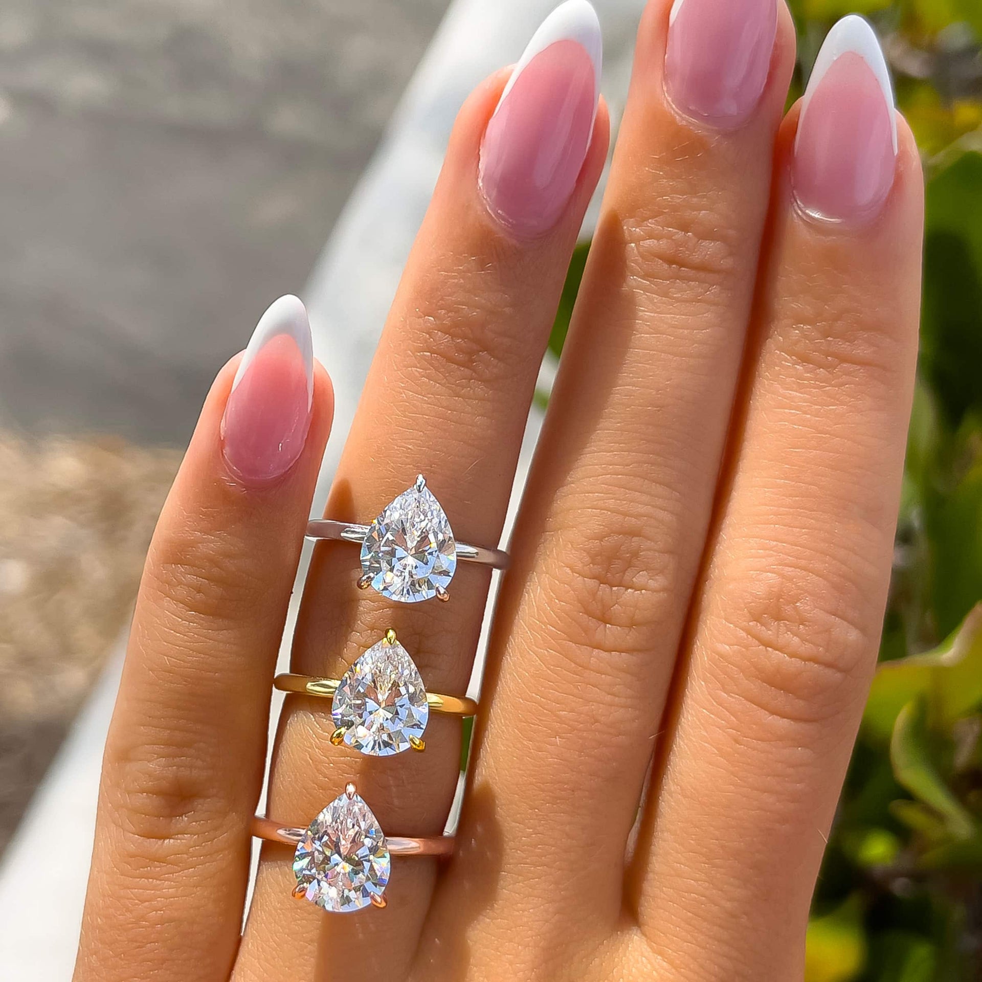 a 3 carat pear cut solitaire engagement ring with a hidden halo shown in silver, gold, and rose gold modeled on a female hand with french tip almond nails