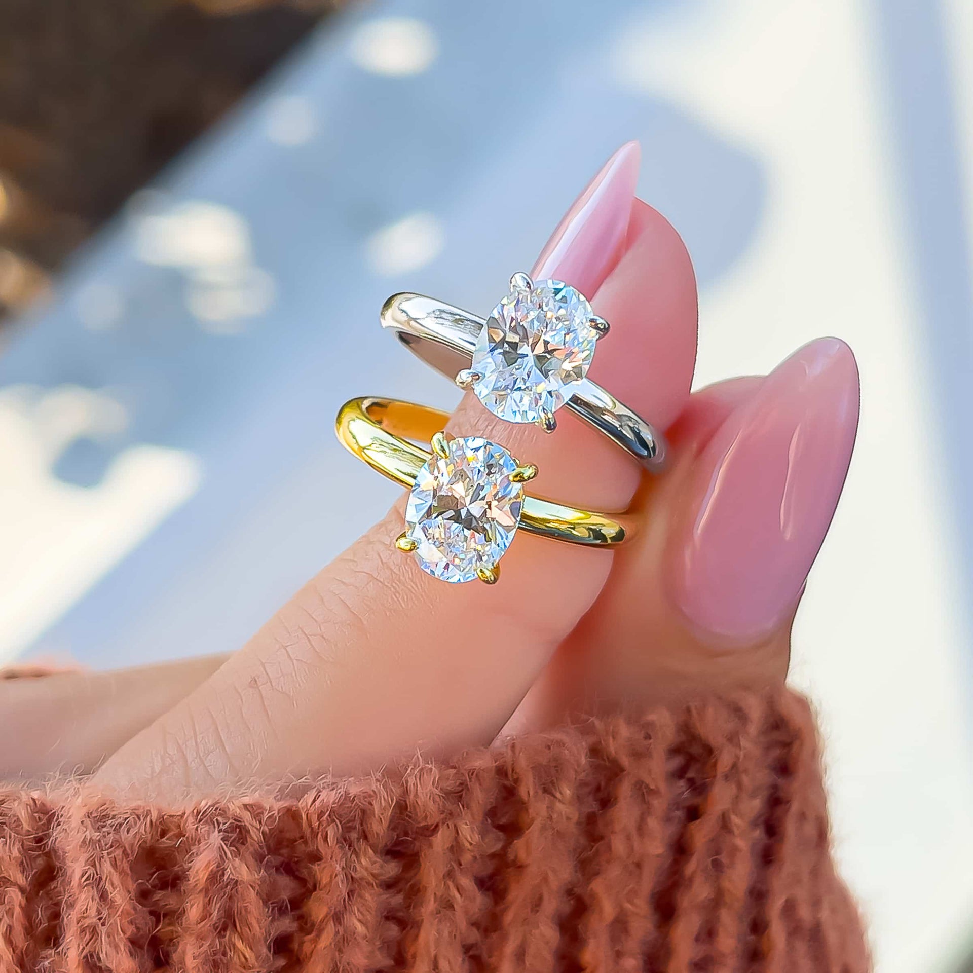 gold and silver engagement rings with shining oval stone on model's finger with pink nails and orange sleeve